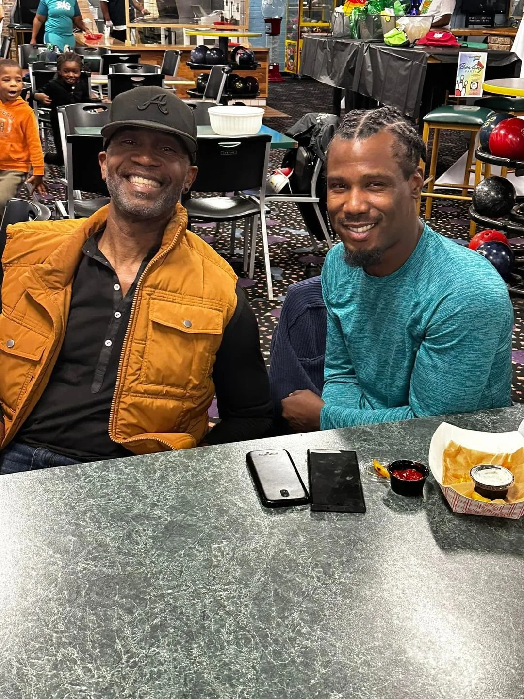 Two smiling men seated at a bowling alley table with food and phones, with children playing in the background and bowling balls on racks nearby.
