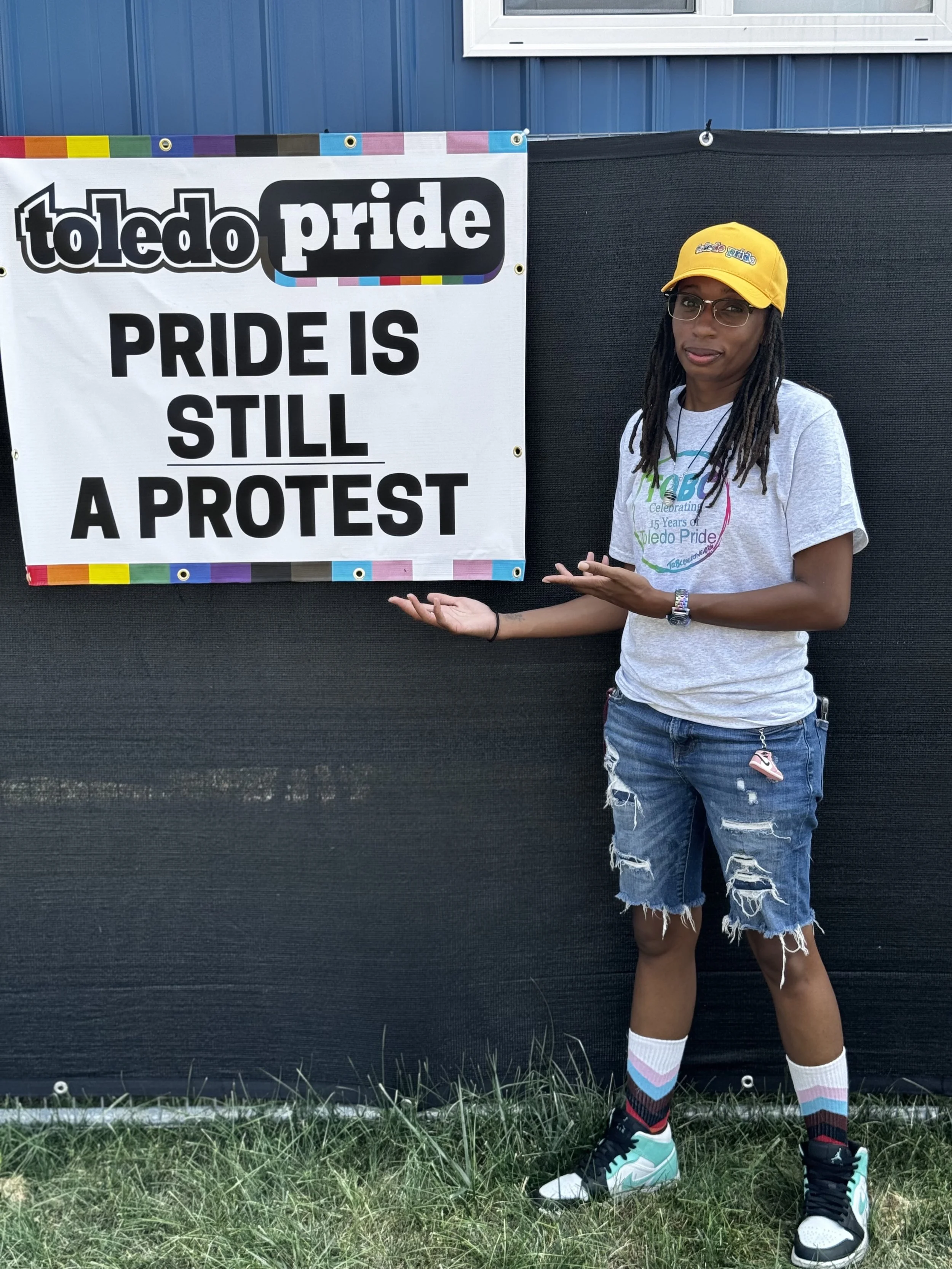 A person standing next to a sign that reads 'Toledo Pride: Pride is still a protest,' wearing a gray T-shirt, distressed denim shorts, colorful socks, and a yellow cap.