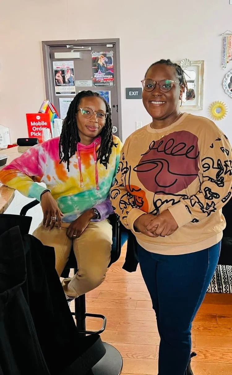 Two women smiling indoors, one sitting on a black stool wearing glasses and a colorful tie-dye hoodie, the other standing nearby wearing glasses and a beige sweatshirt with a red and black graphic design.