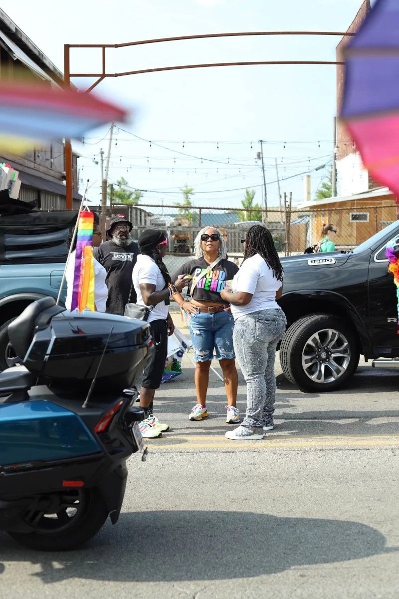 Group of five people standing and talking on a street during a pride parade, with motorcycles and cars decorated with rainbow pride flags and banners in the background.