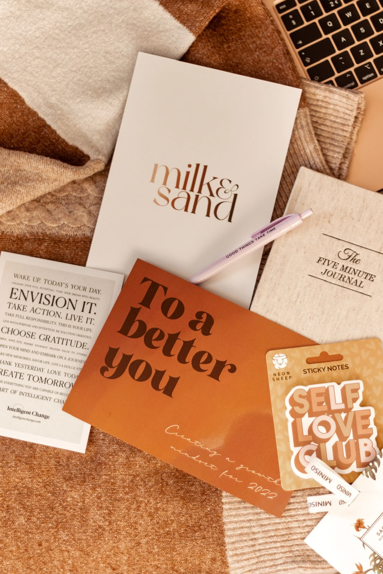 A flat lay of inspirational and motivational items on a cozy fabric surface, including a book titled 'milk & sand', a card that reads 'To a better you', a journal titled 'The Five Minute Journal', a list of positive affirmations, and sticker notes reading 'Self Love Club' with a sheep illustration.