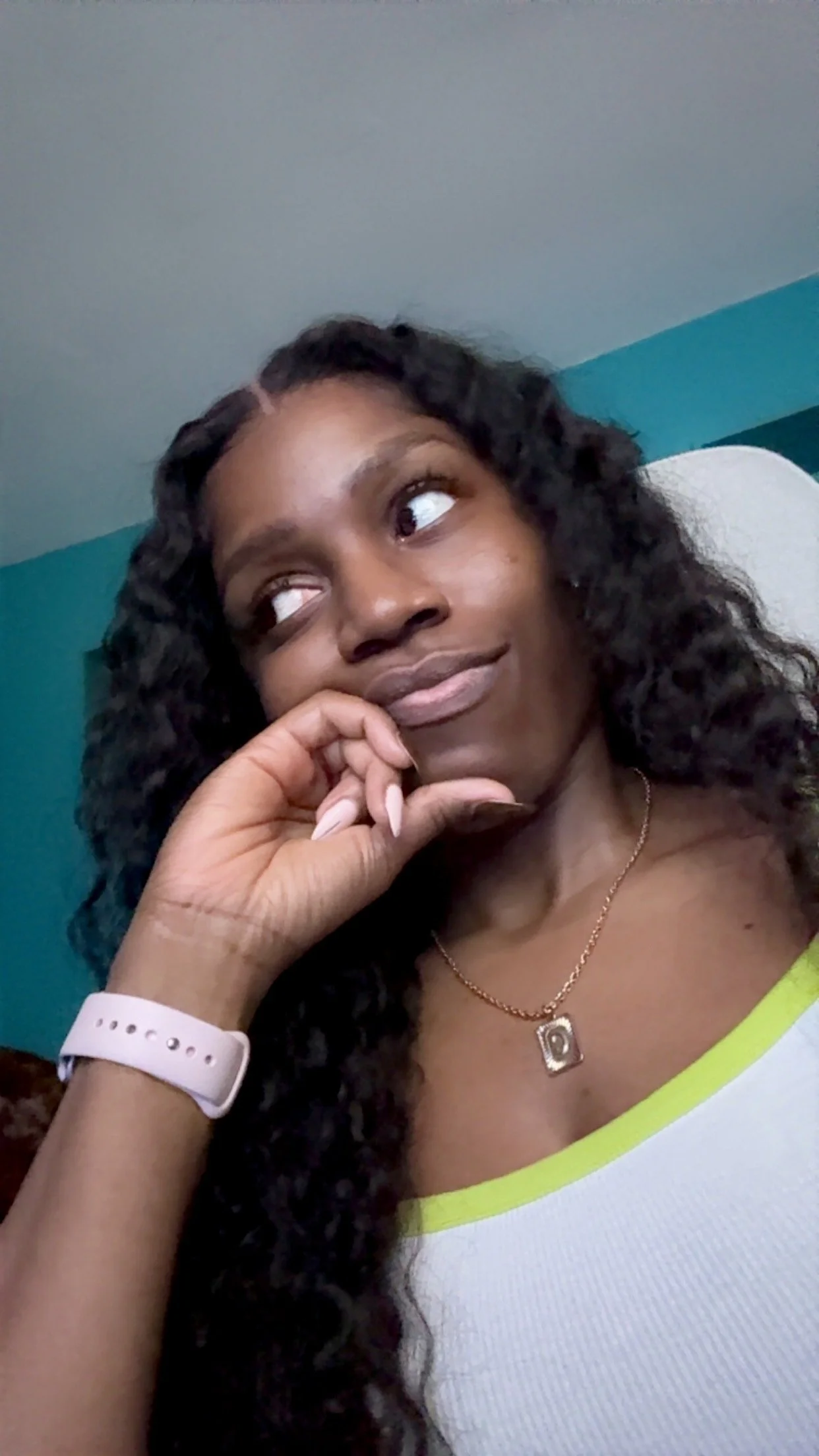 A woman with curly hair sitting on a chair, wearing a white top with lime green trim, a pink watch, and a gold necklace, resting her chin on her hand and looking contemplatively to her left.