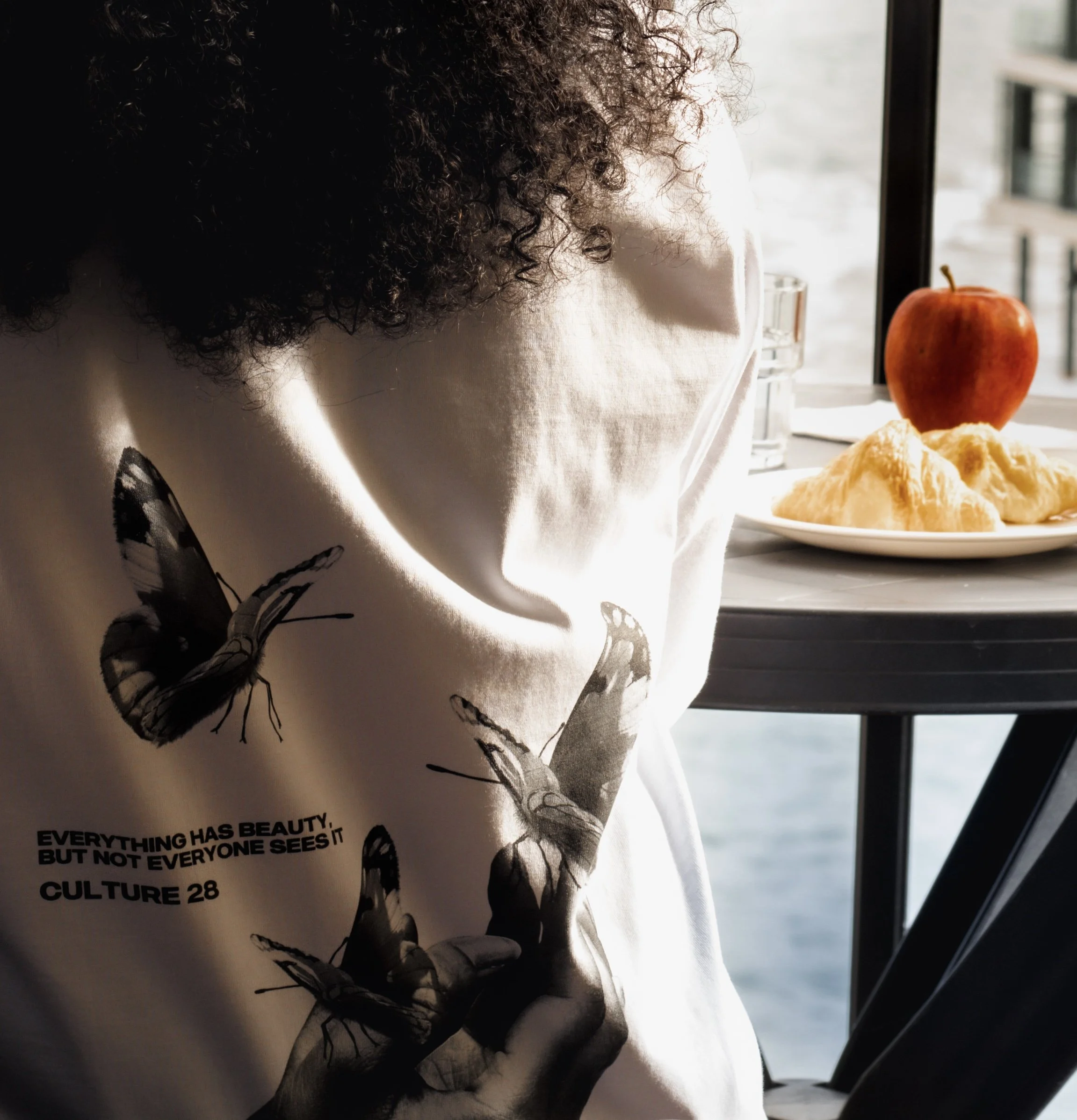 A person with curly hair sitting at a window with a plate of croissants and an apple on the table.