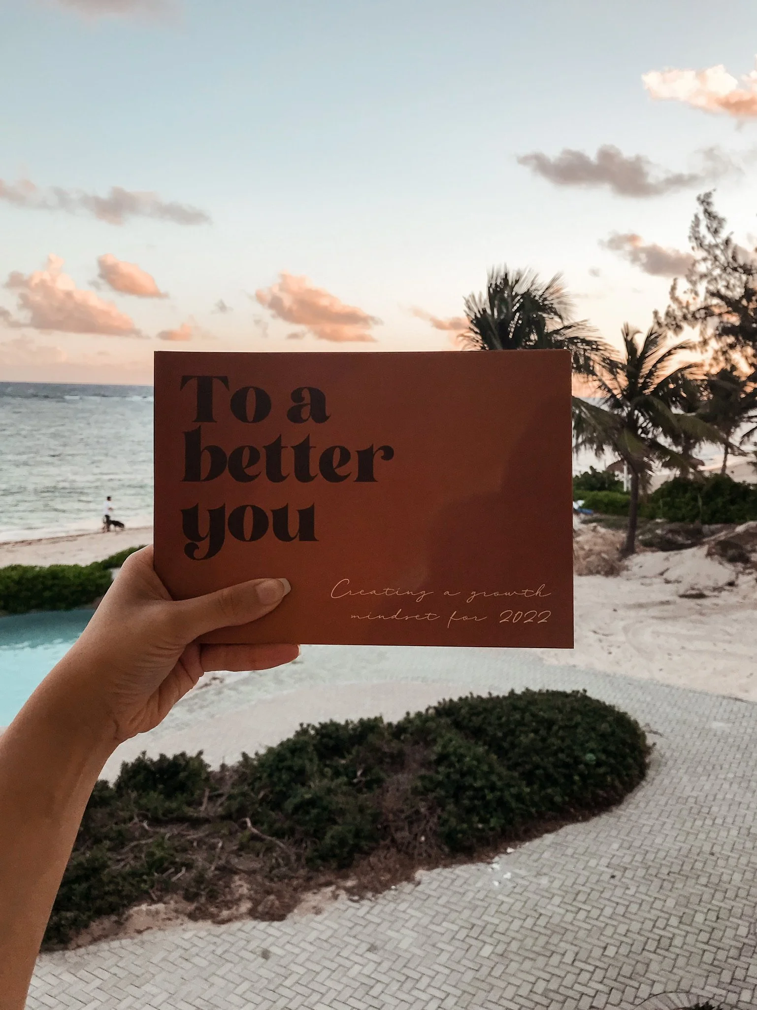 A person holding a motivational card in front of a beach scene at sunset. The card reads 'To a better you. Creating a growth mindset for 2022'. The background features palm trees, sandy beach, ocean, and a pink, orange, and blue sky with clouds.