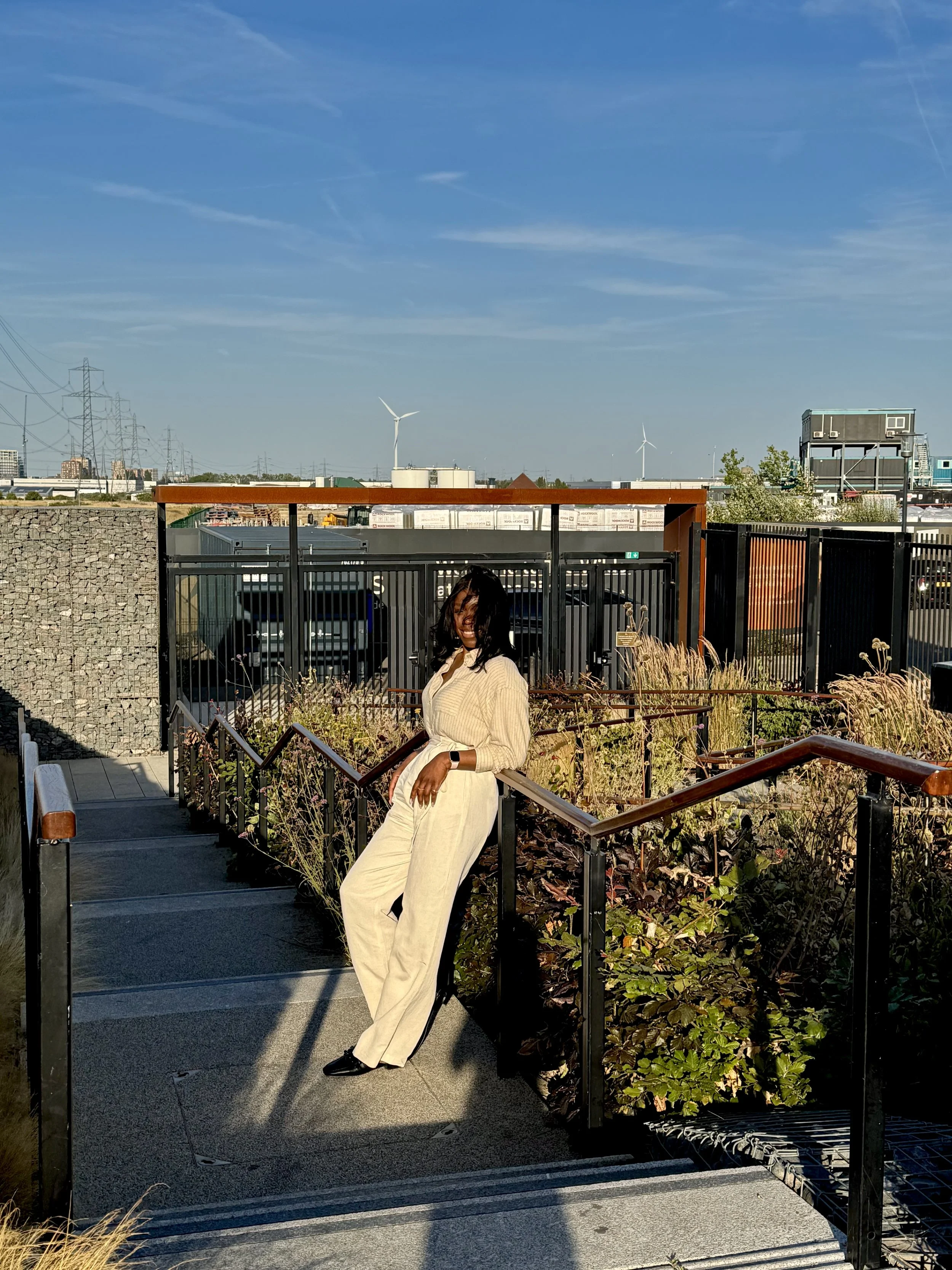 A woman wearing beige pants and a beige top standing on an outdoor walkway, leaning against a black and wooden railing, with a cityscape, wind turbines, and a blue sky in the background.