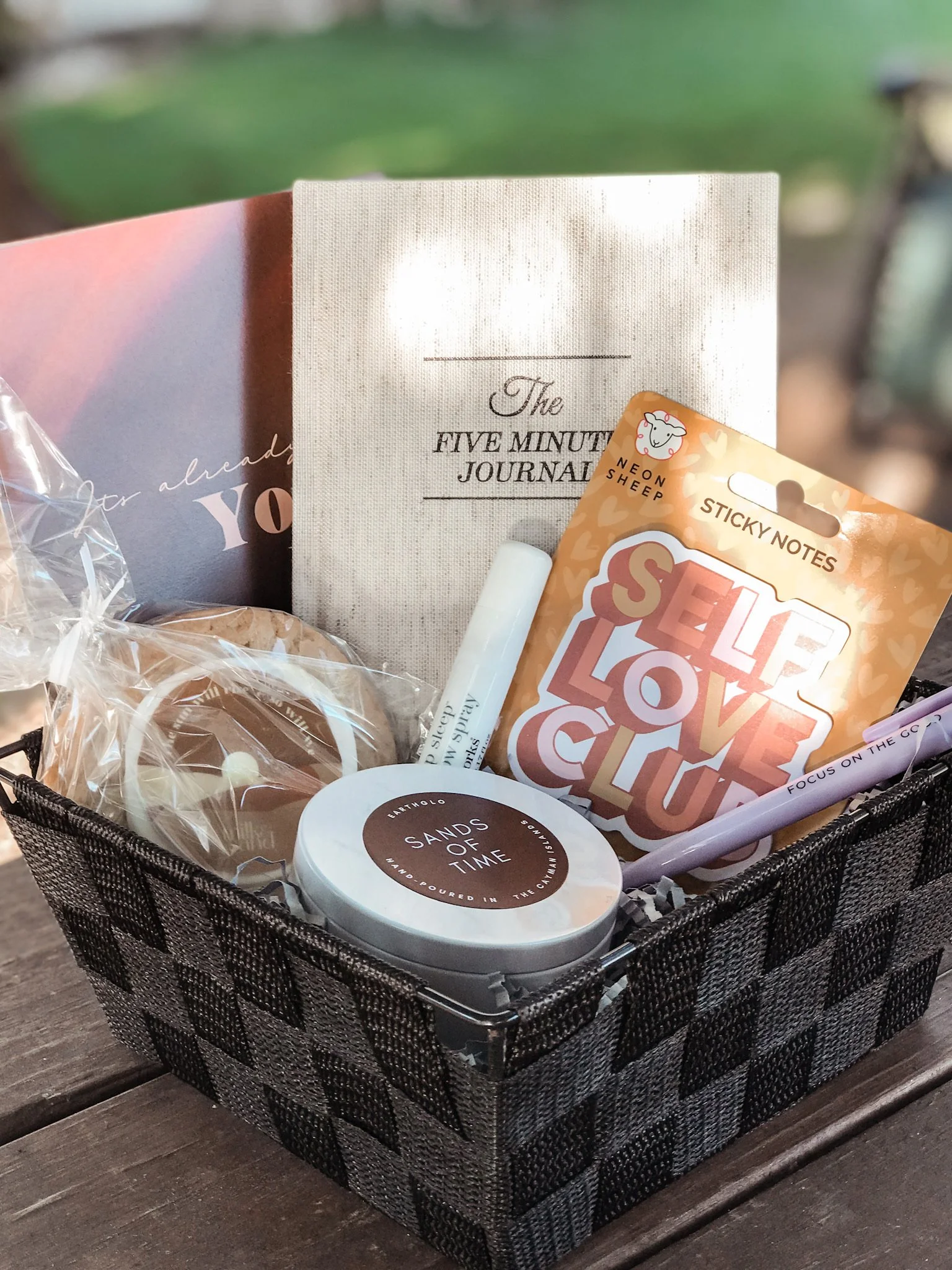 A small black and gray checkered basket containing various items including a white multi-round candle labeled 'SANDS OF TIME', a white lip spray, a purple pen, a sticky notes pad with 'SELF LOVE' printed on it, and a beige notebook titled 'The FIVE MINUTE JOURNAL'.