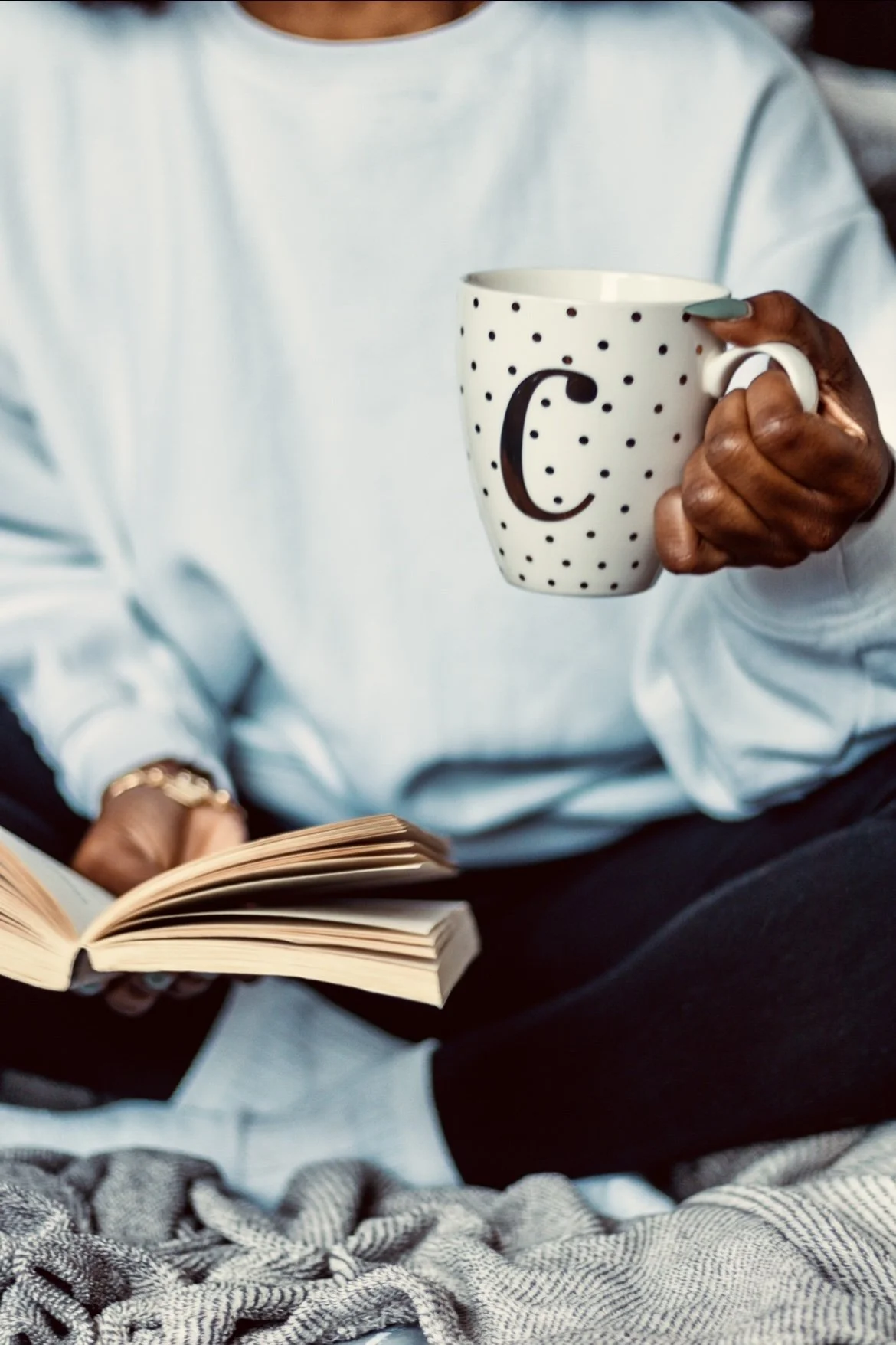 Close-up of a person holding a white mug with black polka dots and a letter 'C' on it, while sitting with an open book.