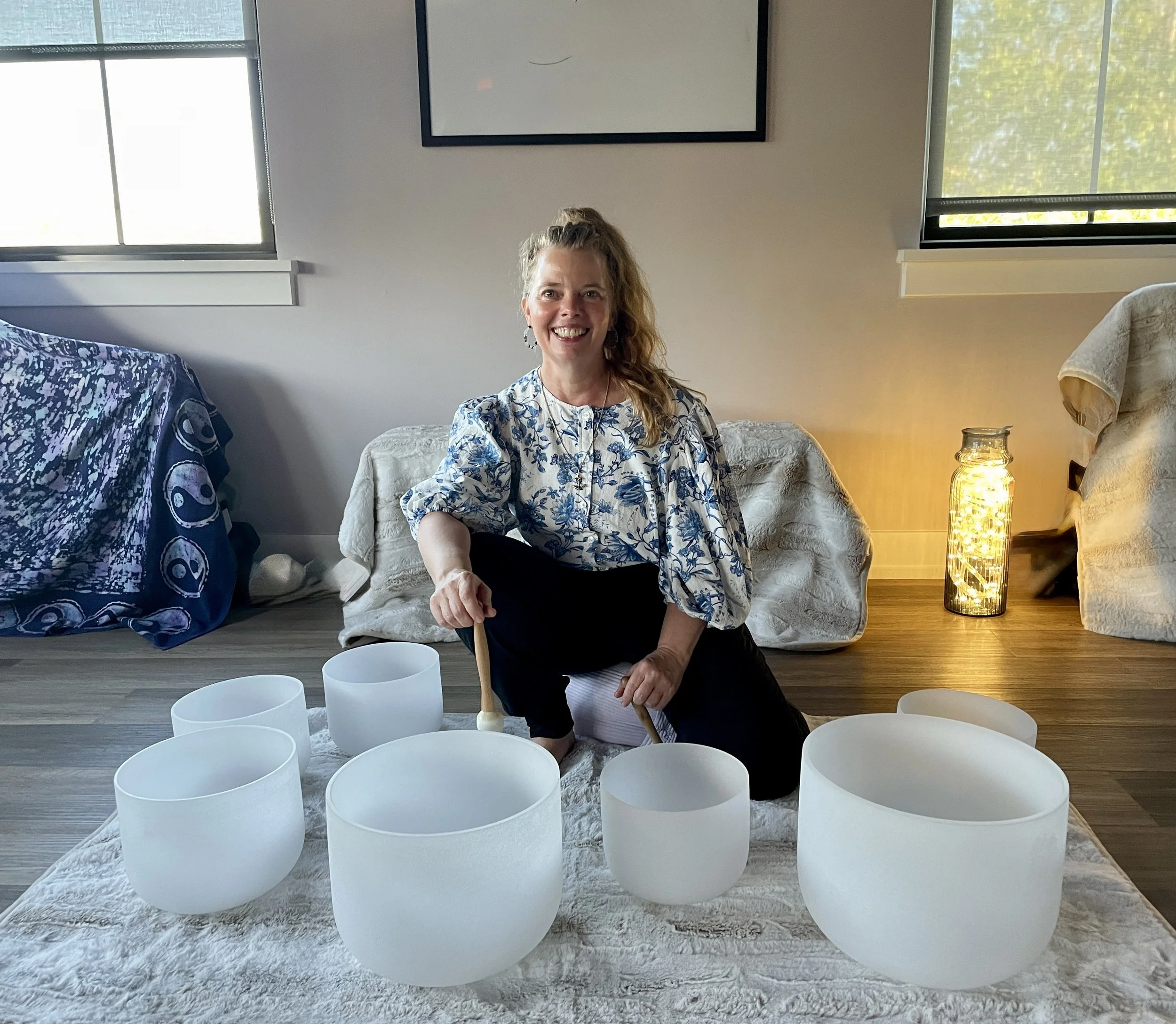 A woman sitting on a white couch in a living room, surrounded by white singing bowls, with a wall-mounted TV, windows letting in natural light, and a candle on a side table.