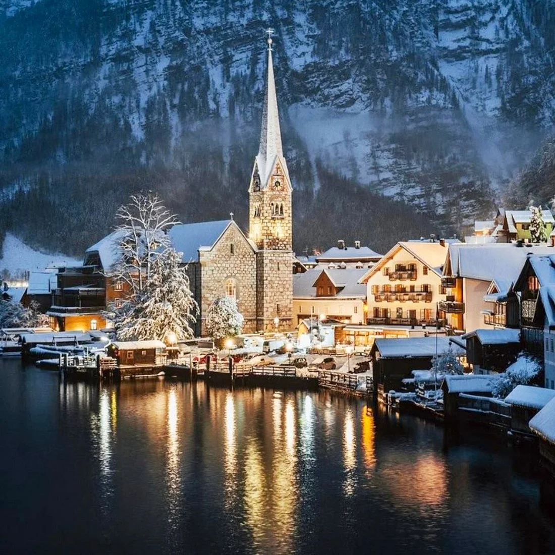 An illuminated church, Parish of the Assumption, amongst snow-capped buildings. Snowy mountains are in the foreground of the image