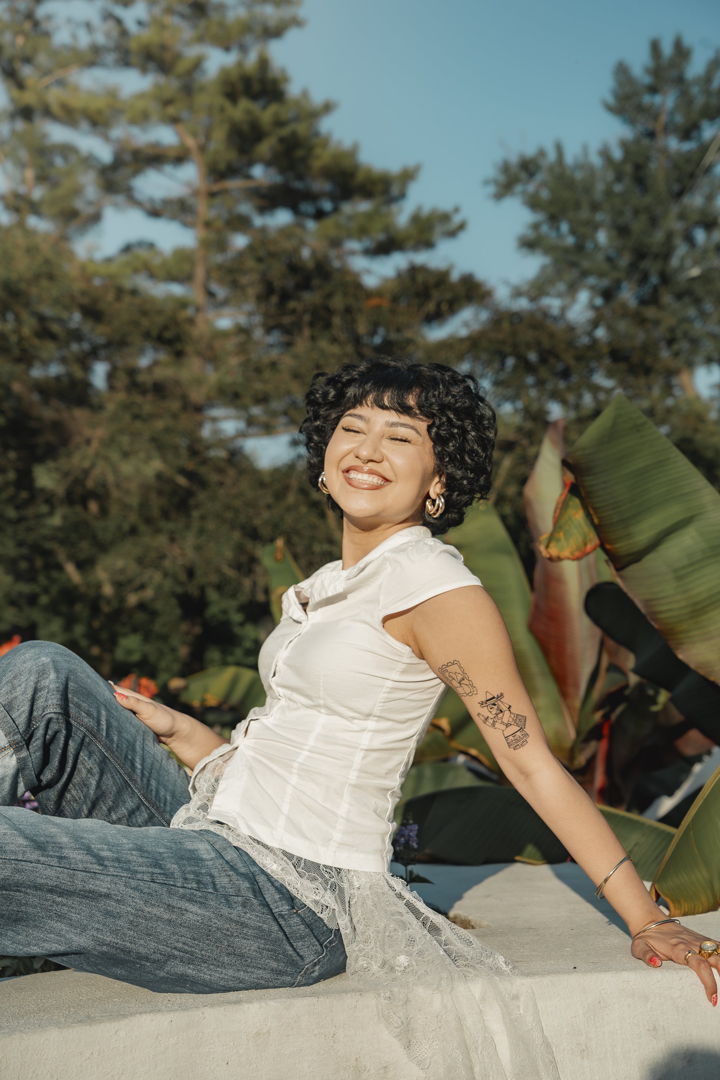 A young woman with short curly black hair and tattoos on her arm, smiling with her eyes closed, sitting outdoors among large green leaves and trees during daytime.