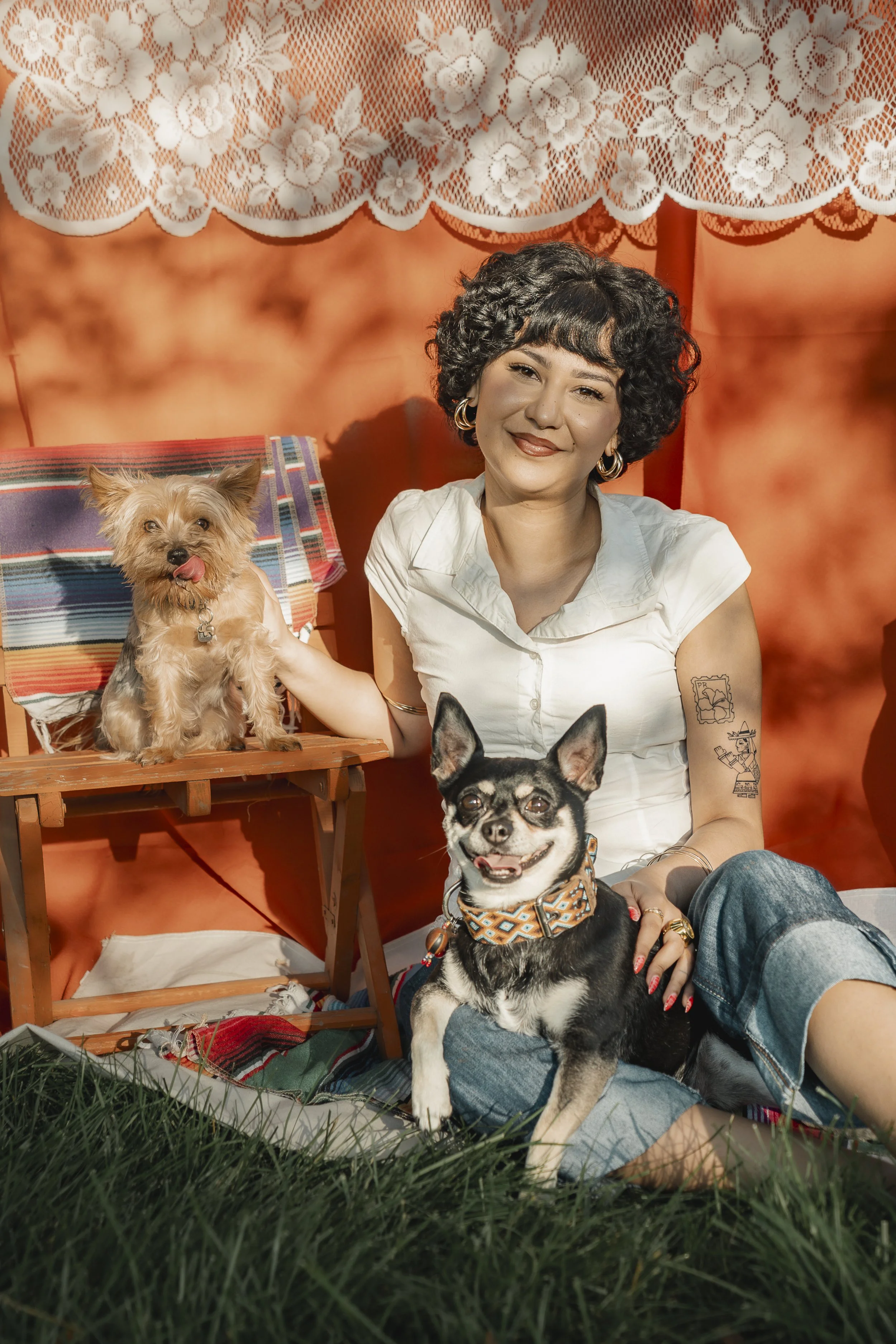 A woman with short curly black hair, wearing a white shirt and jeans, sitting on grass with two dogs, a small tan dog with its tongue out sitting on a small wooden bench, and a black and white dog with a patterned scarf around its neck, sitting on her lap. The background features a large orange wall with lace curtains hanging above.