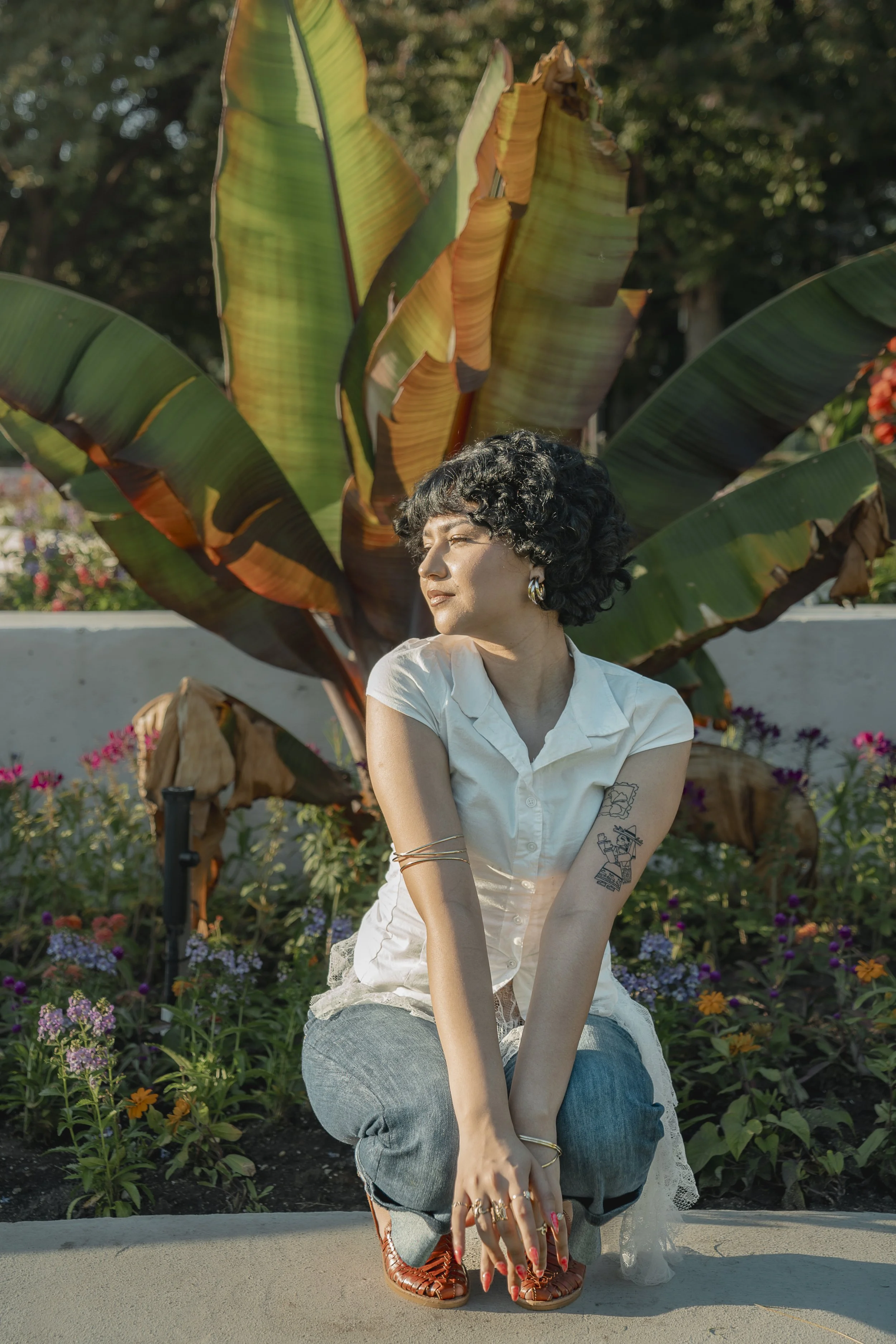 A woman with short curly black hair and tattoos on her arms posing outdoors in front of large green and yellow banana leaves, surrounded by colorful flowers, with sunlight on her face.