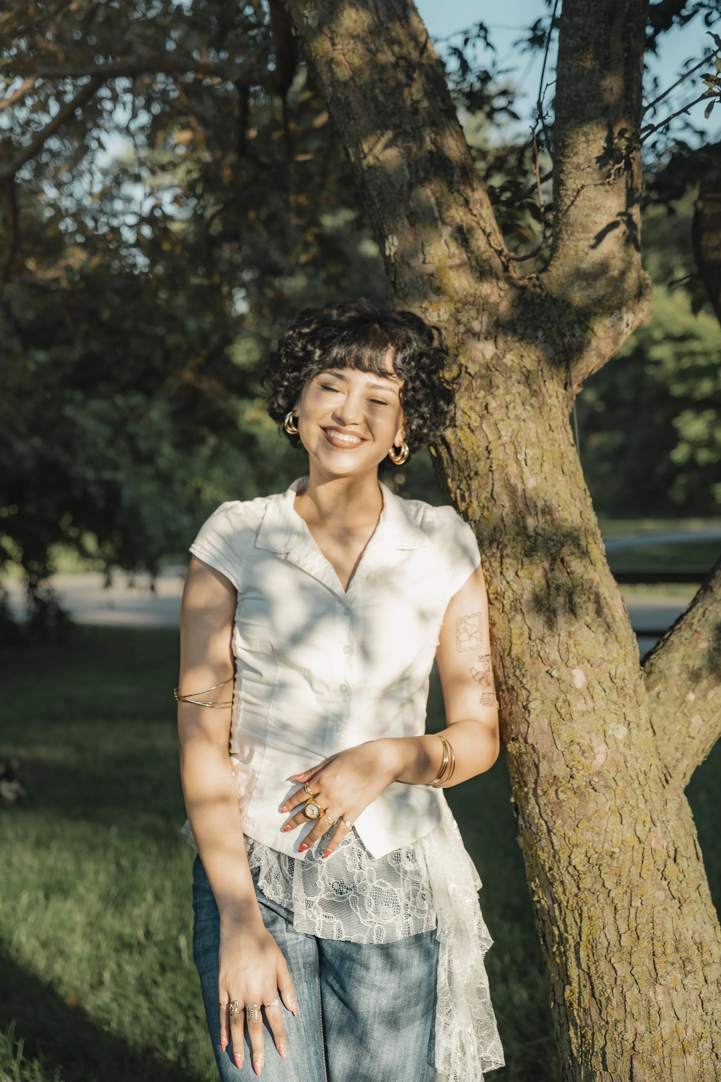 A woman with curly short hair smiling and standing outdoors by a tree, wearing a white short-sleeve shirt, jewelry, and lace detail arm.