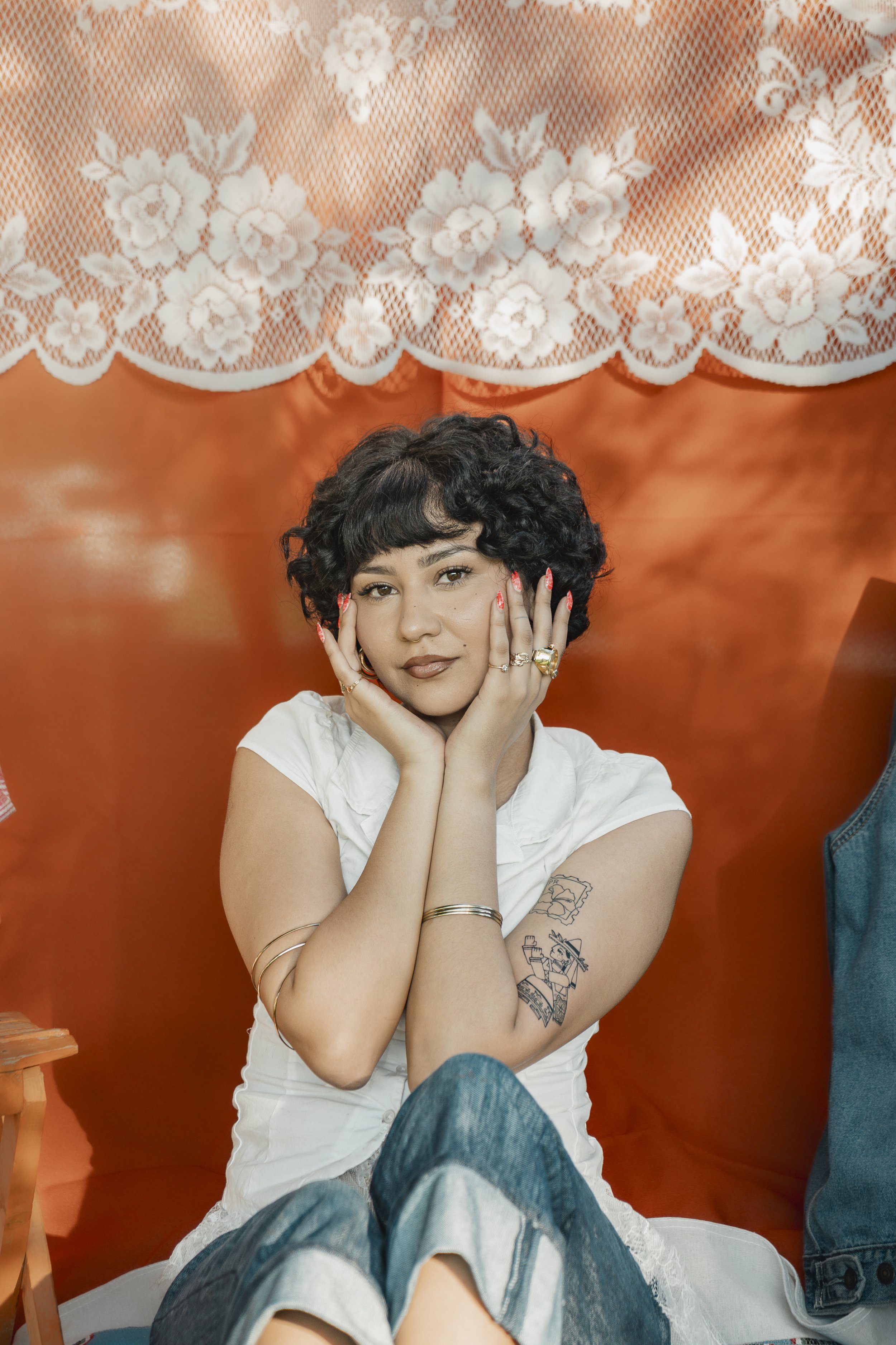 A woman with dark, curly hair and tattoos on her arm, wearing a white top and multiple rings, sits against an orange-colored background and rests her head on her hands.