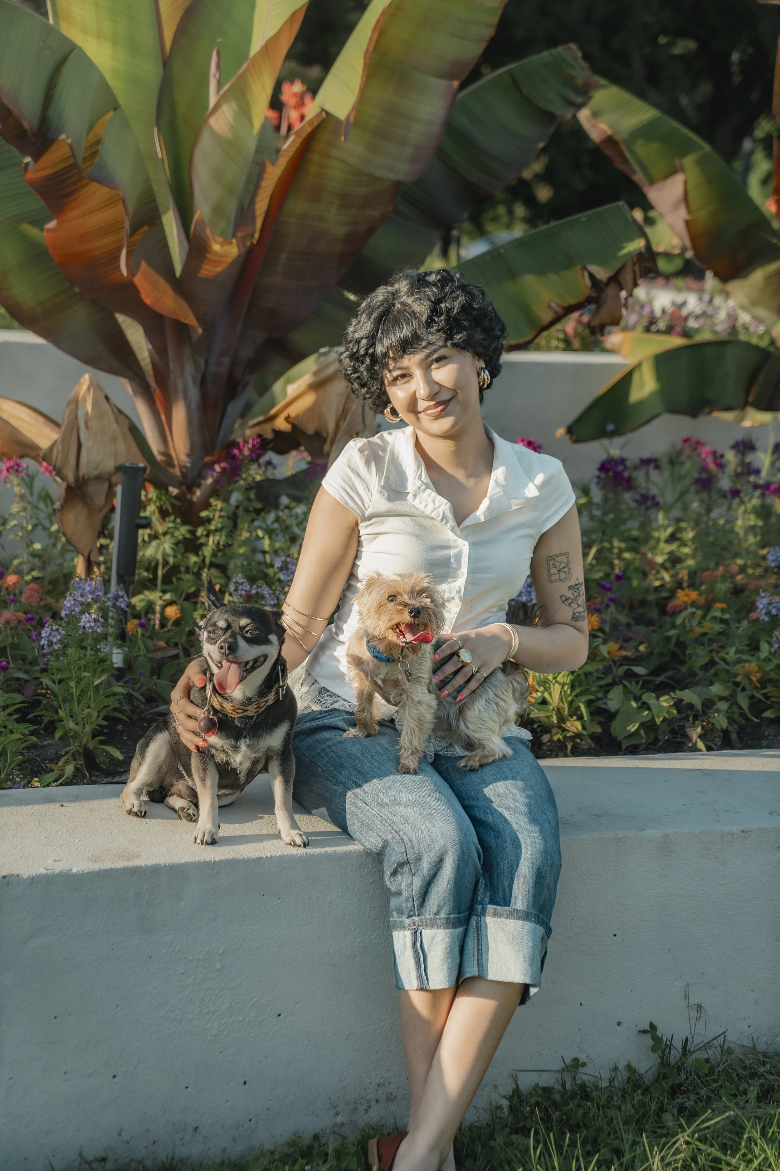 A woman with short curly hair wearing a white shirt and denim shorts sitting outdoors on a concrete ledge, holding two small dogs, with large green leaves and colorful flowers in the background.