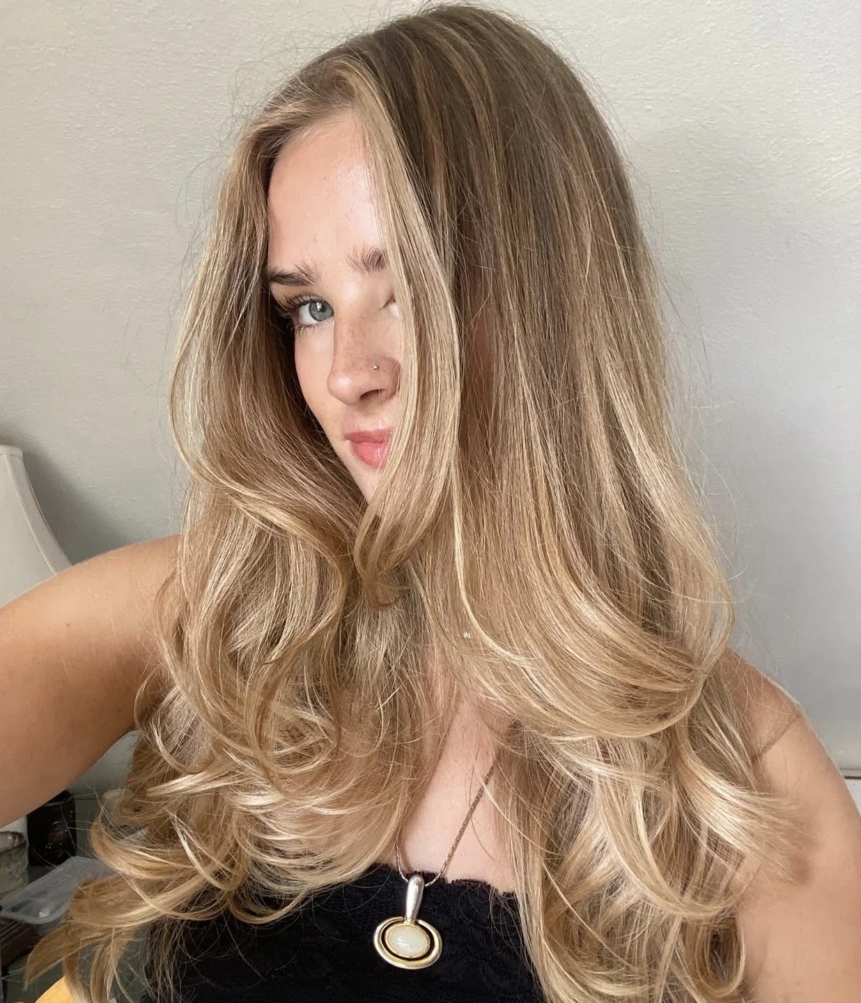 A woman with long, voluminous blonde hair, blue eyes, and a nose piercing, taking a selfie in front of a neutral wall.