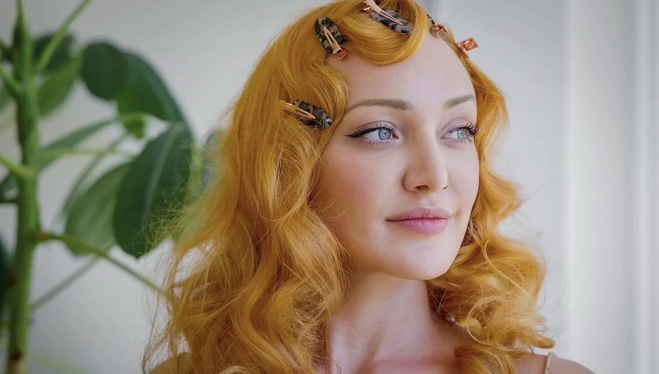 A woman with curly red hair and blue eyes looks thoughtfully to the side, with hair clips in her hair, standing near a green plant indoors.