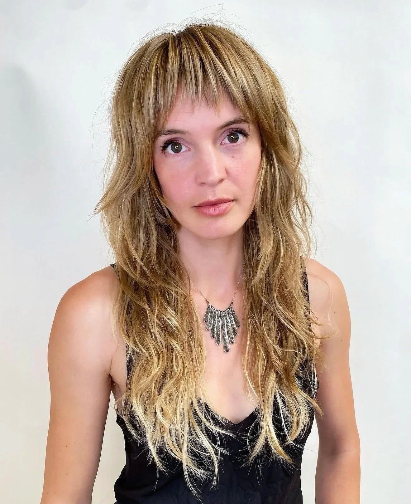 A woman with long, wavy blonde hair and light skin, wearing a black sleeveless top and a silver necklace, standing against a plain white background.
