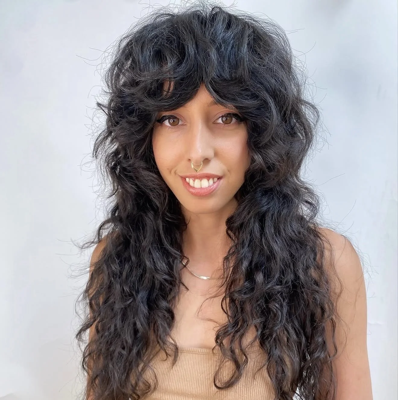 A woman with long, curly black hair and light brown eyes, smiling, wearing a beige top, a small necklace, and a septum piercing, standing against a plain white background.