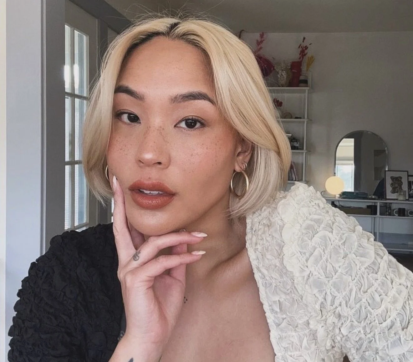 A young woman with blonde hair, brown eyes, freckles, and hoop earrings looking into the camera with her hand resting on her chin in a well-lit room.