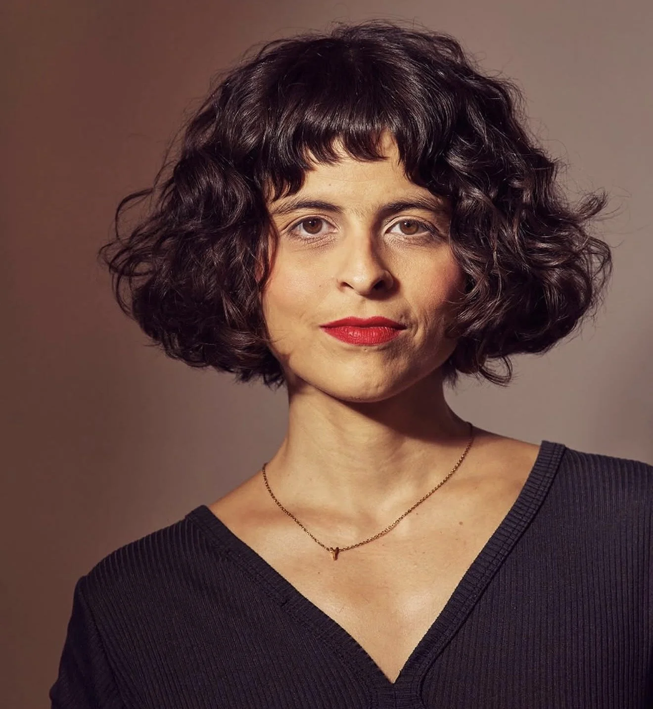 A woman with short curly dark hair, wearing red lipstick, a black V-neck top, a small gold necklace, and a subtle smile, posing against a brown background.