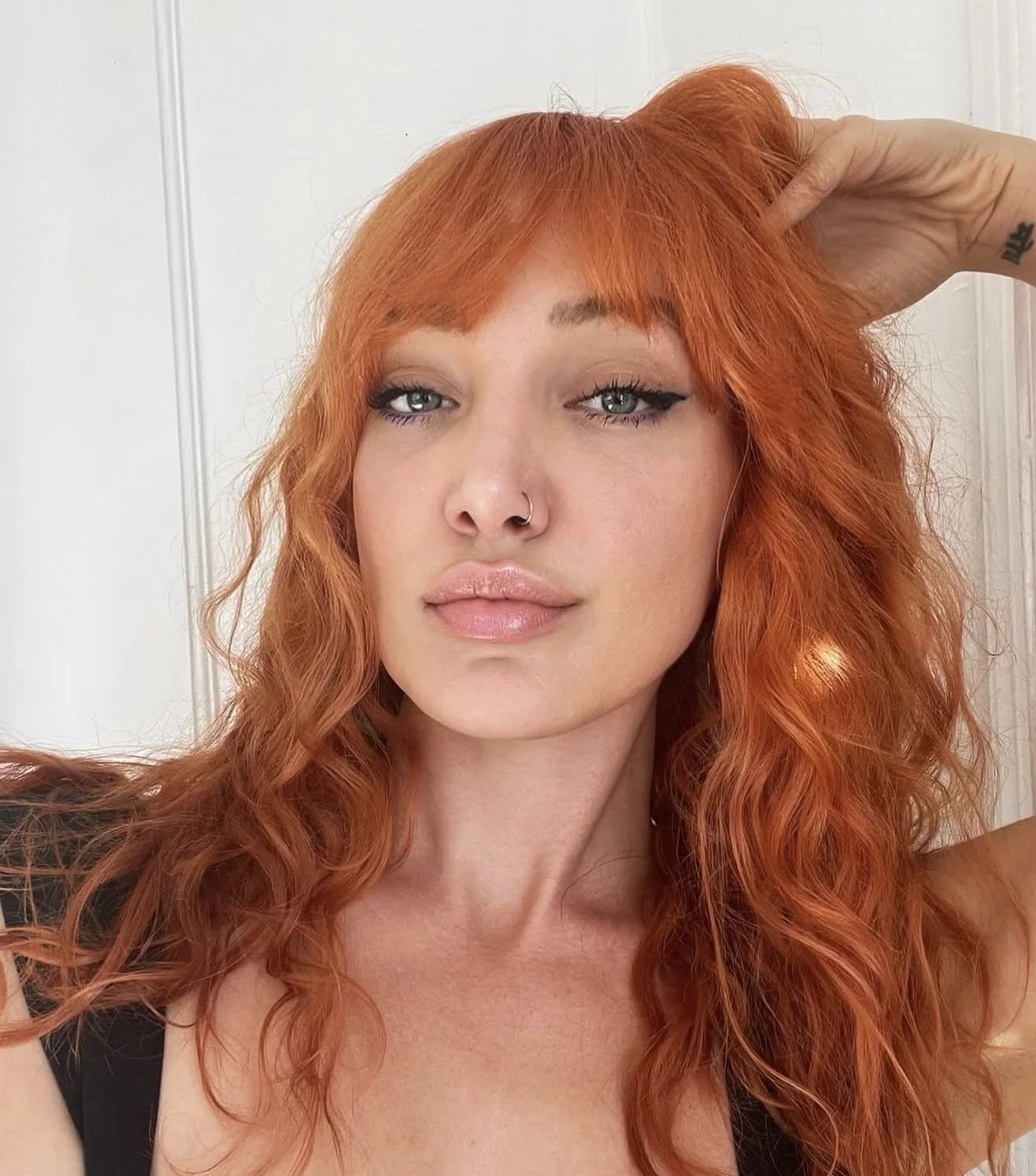 A woman with curly red hair and blue eyes taking a selfie against a white wall, wearing a black top and makeup.