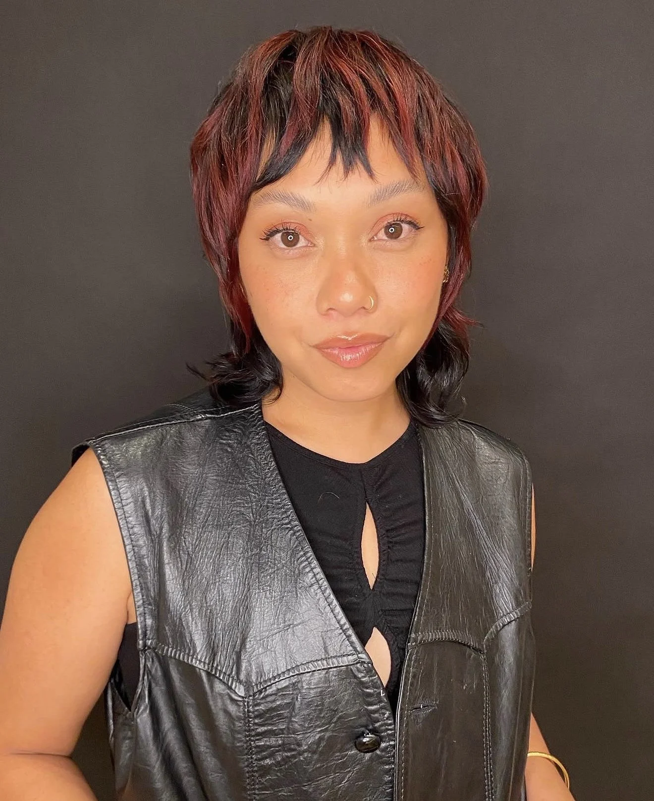 A woman with short, layered, reddish-brown and black hair, wearing a black sleeveless top and a black leather vest, standing against a plain dark background.