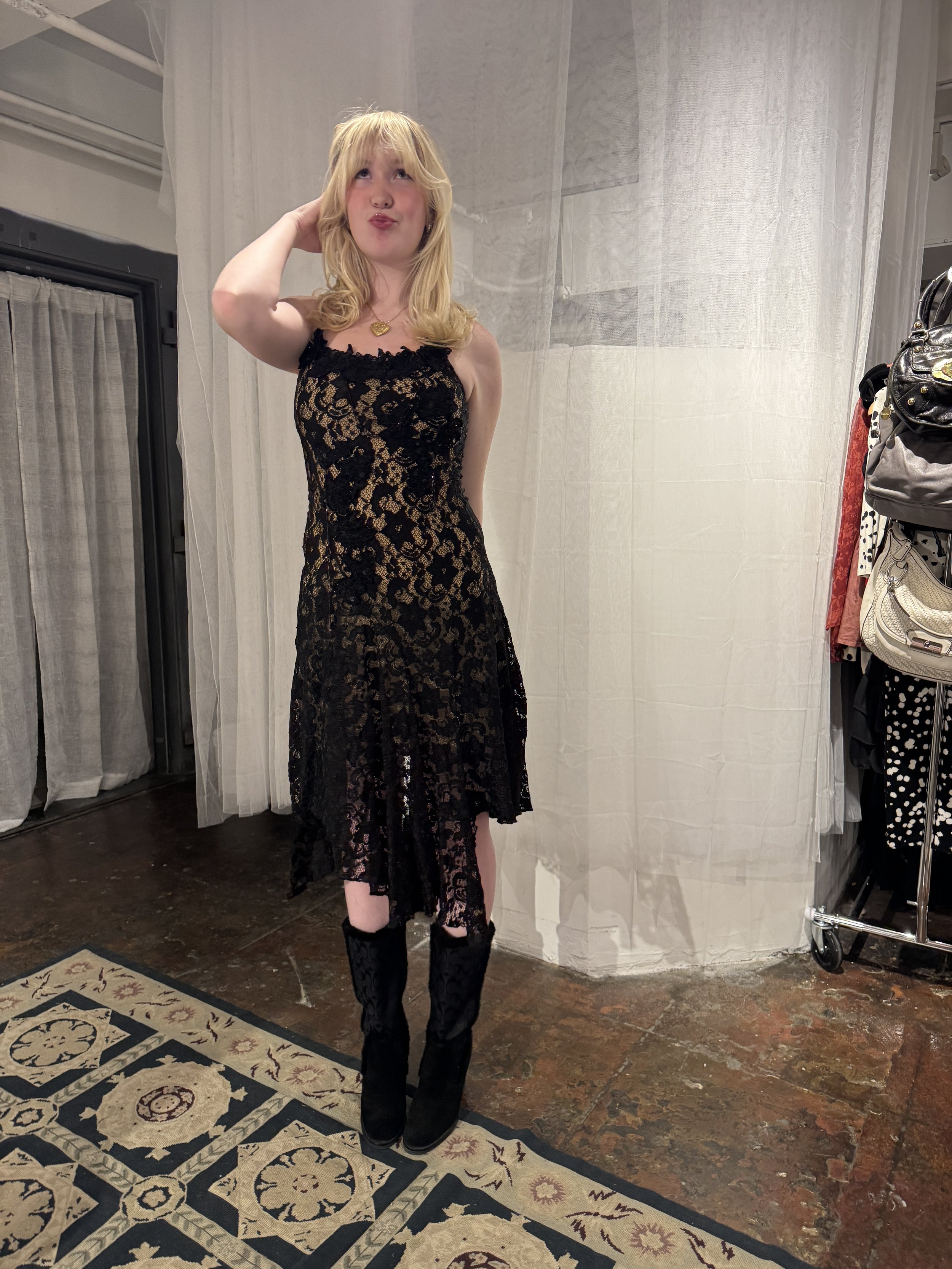 A young woman with blonde hair wearing a black lace dress and tall black boots standing on a patterned rug indoors.