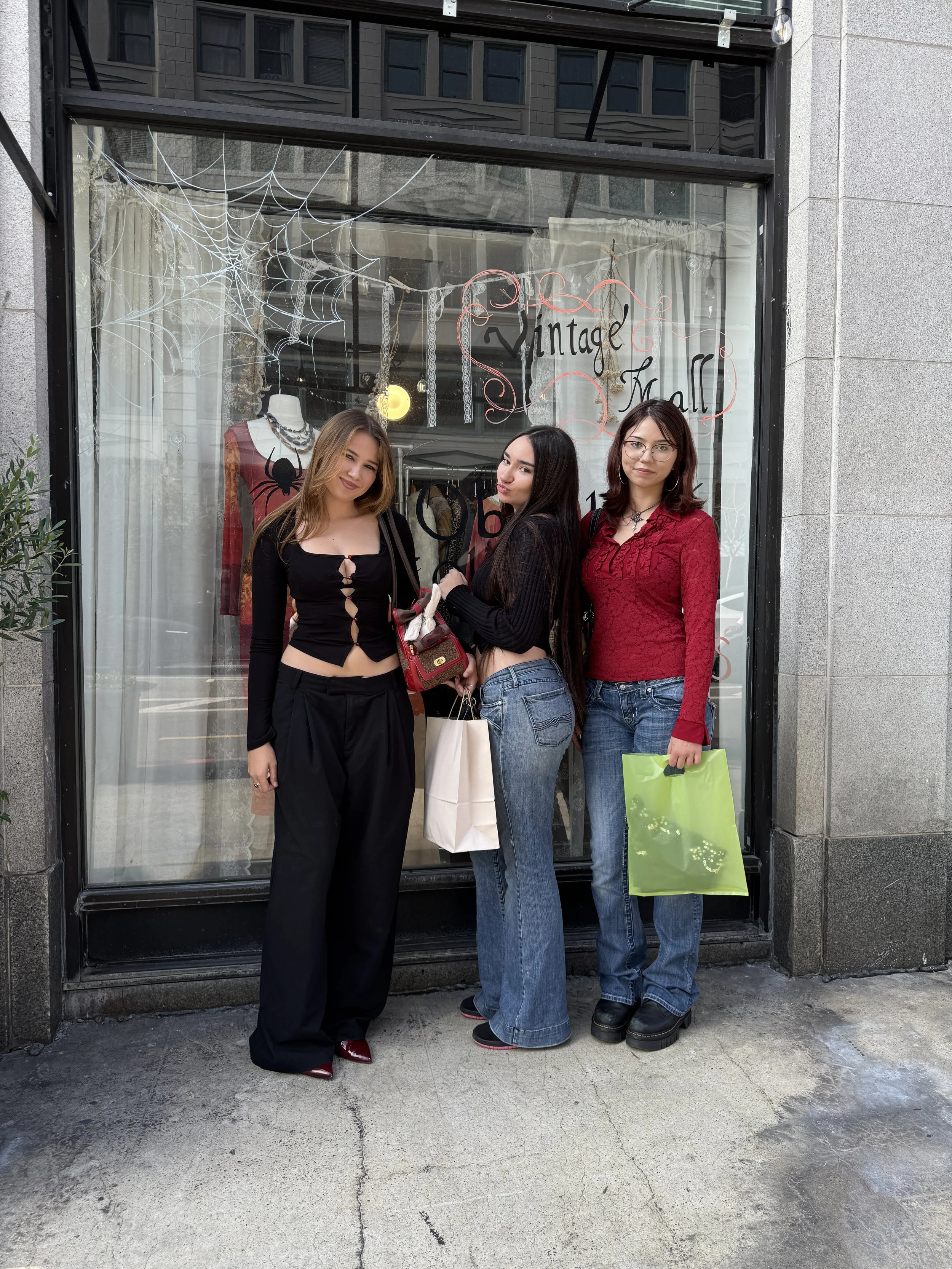 Three young women standing in front of a vintage mall storefront with Halloween decorations, including a spider web. They are holding shopping bags and posing for the photo.