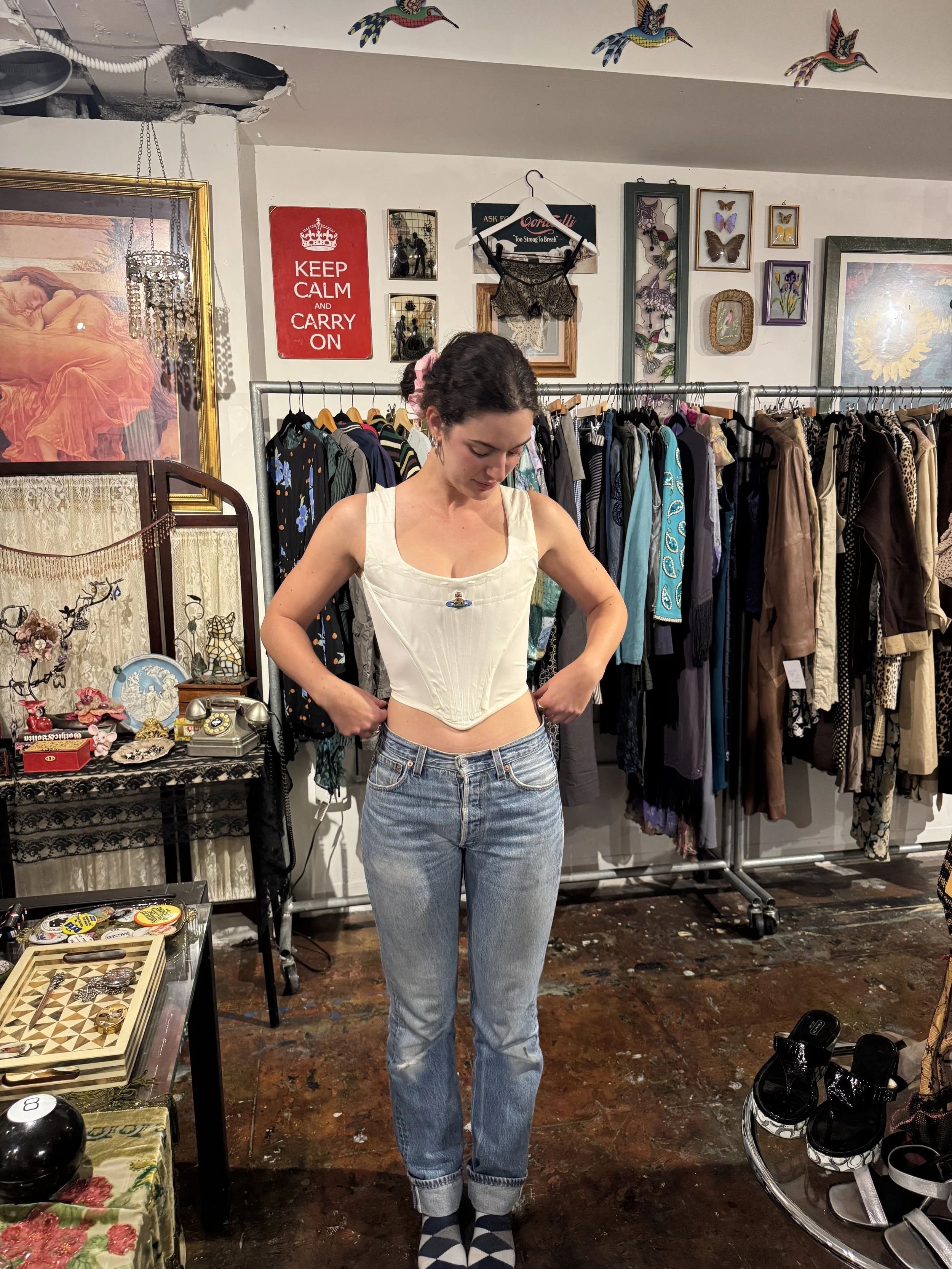 A woman with dark hair, wearing a white corset top and blue jeans, is standing in a vintage clothing store, adjusting her top. The store is decorated with framed art, butterflies, and vintage accessories, with clothing racks in the background and var
