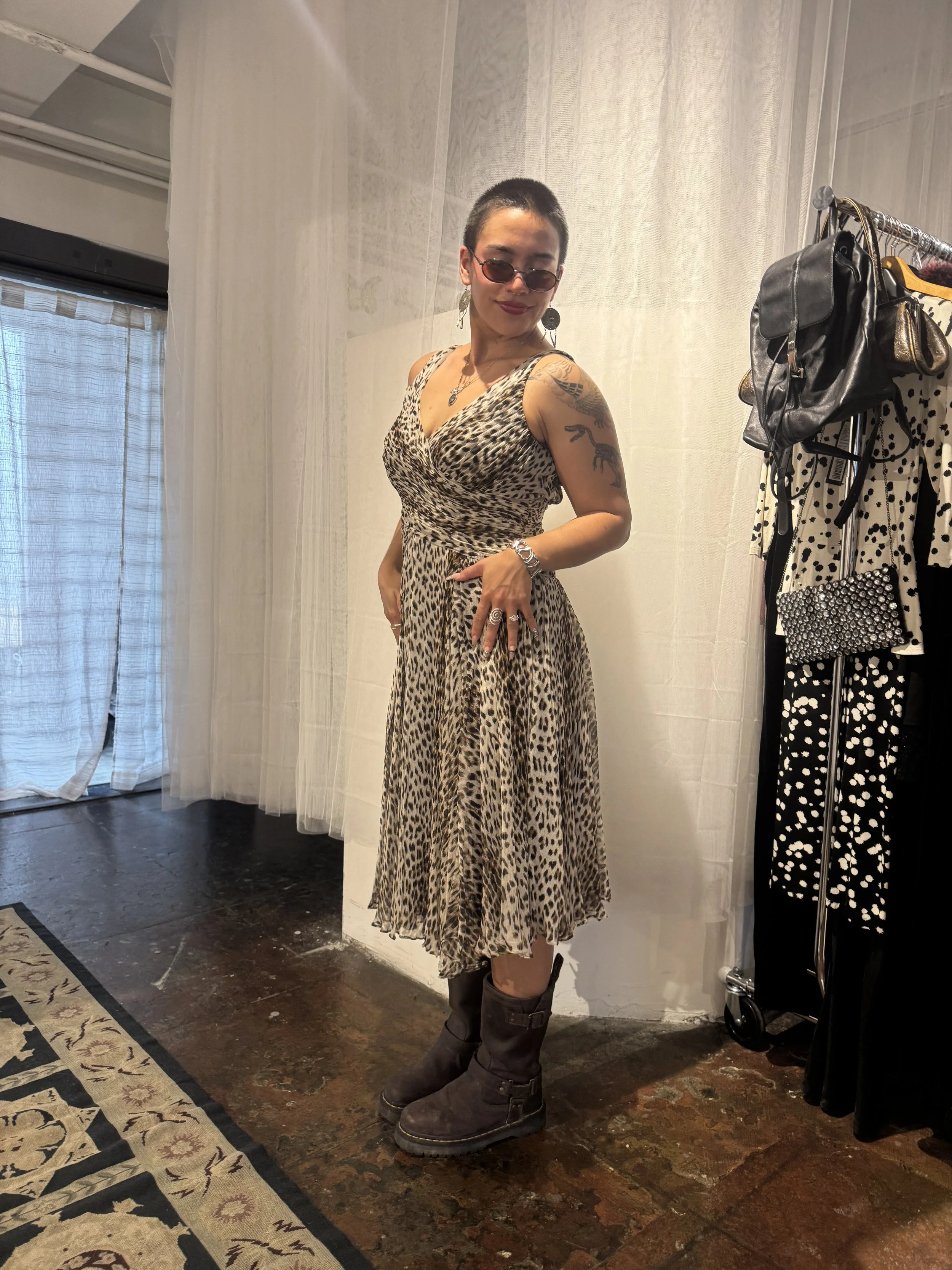 Woman with short hair wearing sunglasses, a leopard print dress, and boots, standing indoors next to a clothing rack.