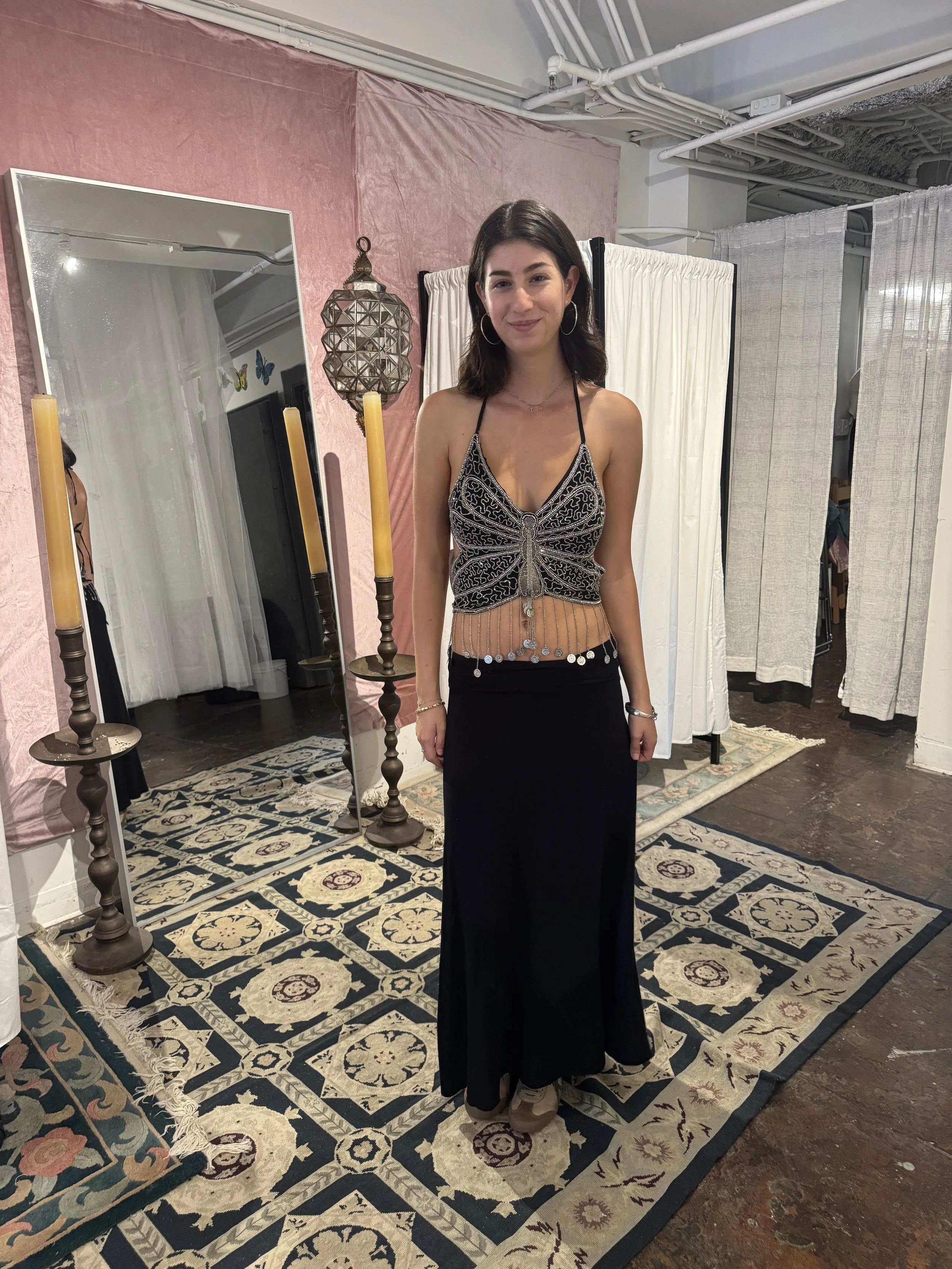 A young woman with dark hair and hoop earrings, wearing a black and silver patterned crop top and a long black skirt, stands indoors in front of a mirror, candles, and a folding room divider.