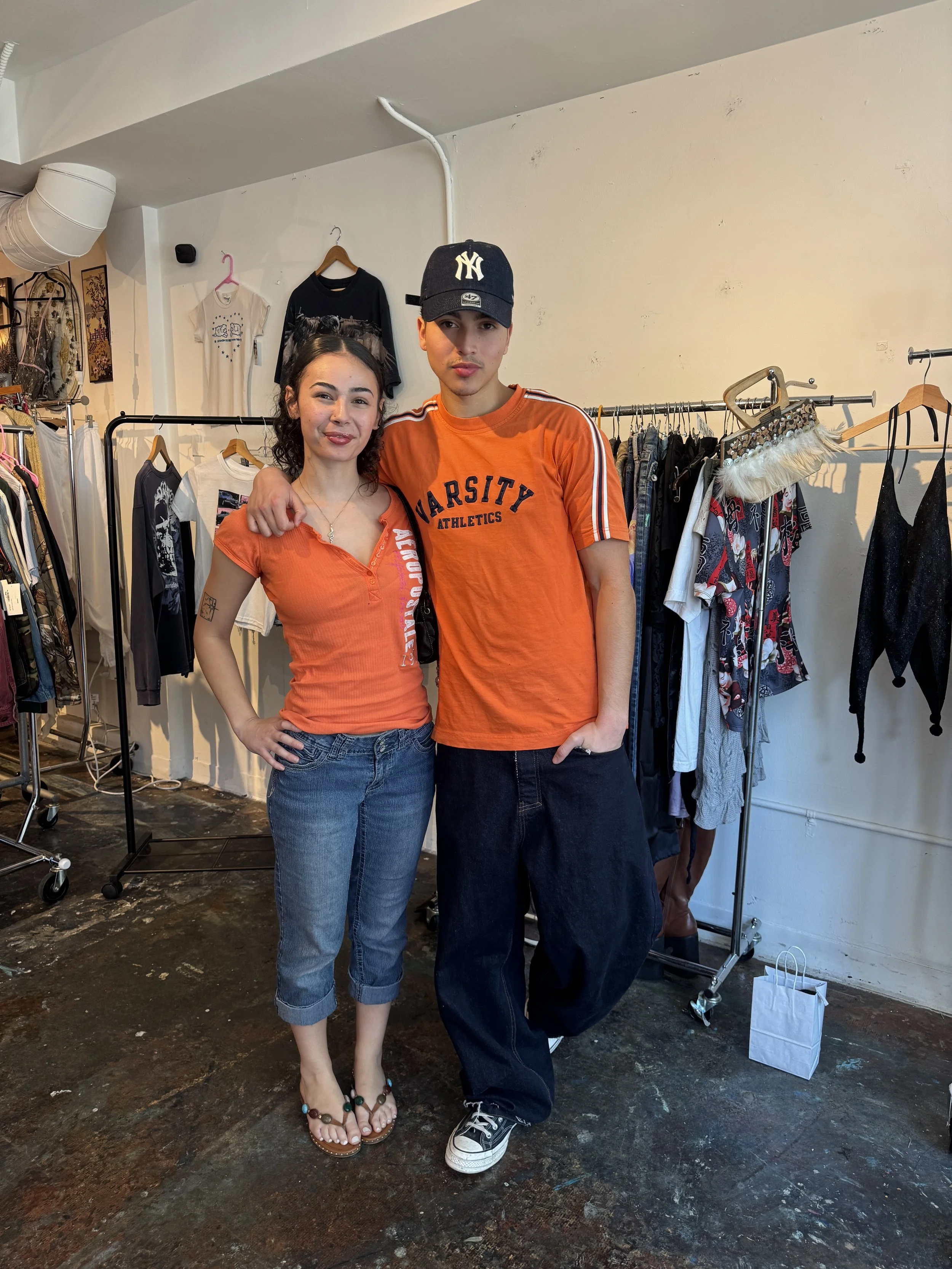 A young man and woman are standing together inside a clothing store, both wearing matching orange athletic shirts. The woman has curly hair, is smiling, and has her hand on her hip. The man is wearing a New York Yankees cap and has his arm around the