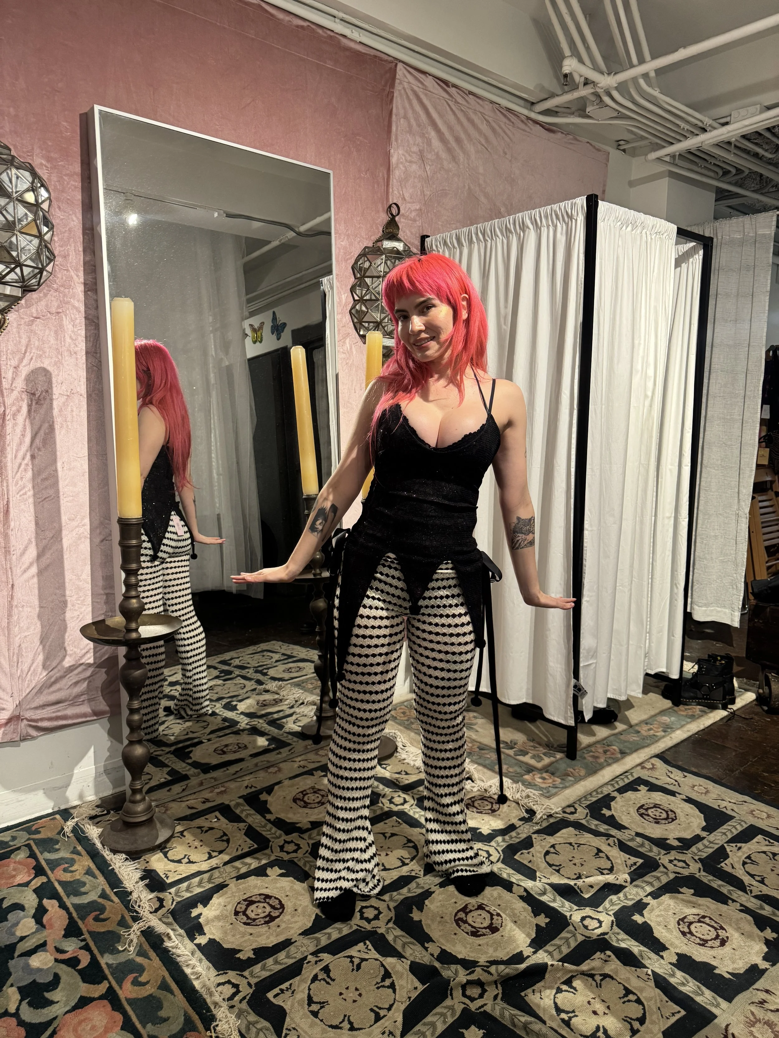 A woman with bright pink hair and tattoos, wearing a black tank top and black-and-white patterned pants, standing in front of a mirror, with a fabric divider to her side and a decorative rug on the floor.