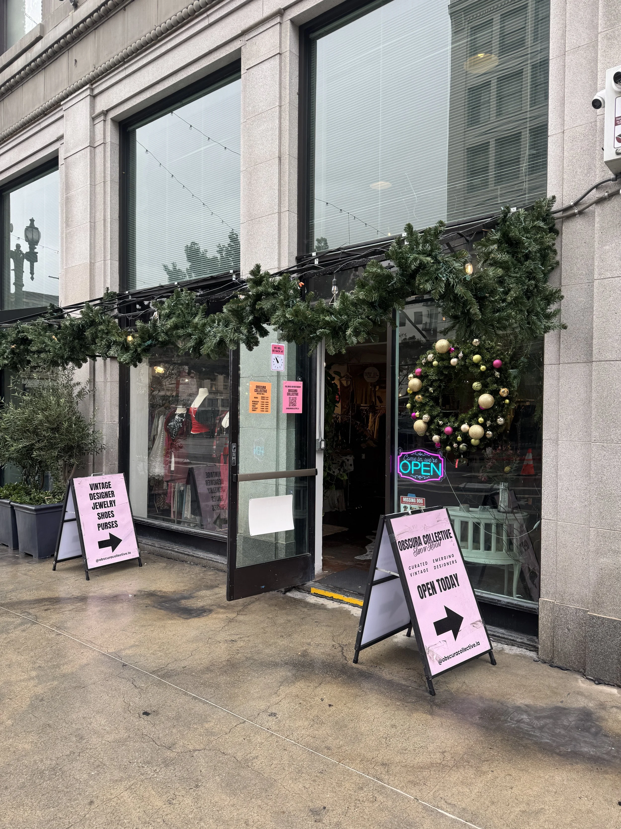Storefront decorated for Christmas with a green garland and wreaths, pink and gold ornaments, and pink signs with arrows indicating it is open today.