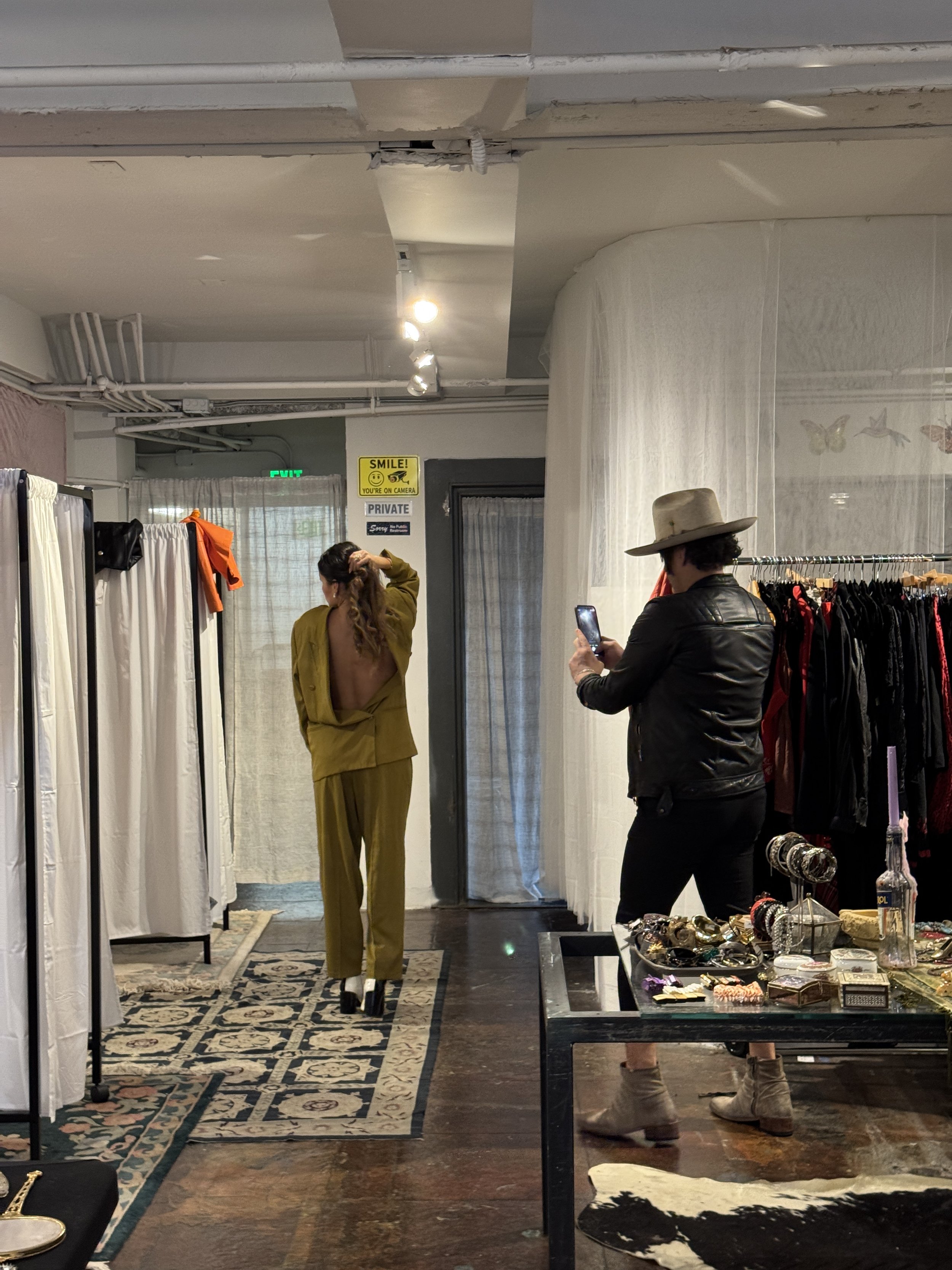 A woman in a mustard-colored suit with her back exposed, standing in a fitting room area, another woman wearing a black leather jacket and beige hat taking a photo with her phone, a display table with jewelry, and clothing racks with red and black ga