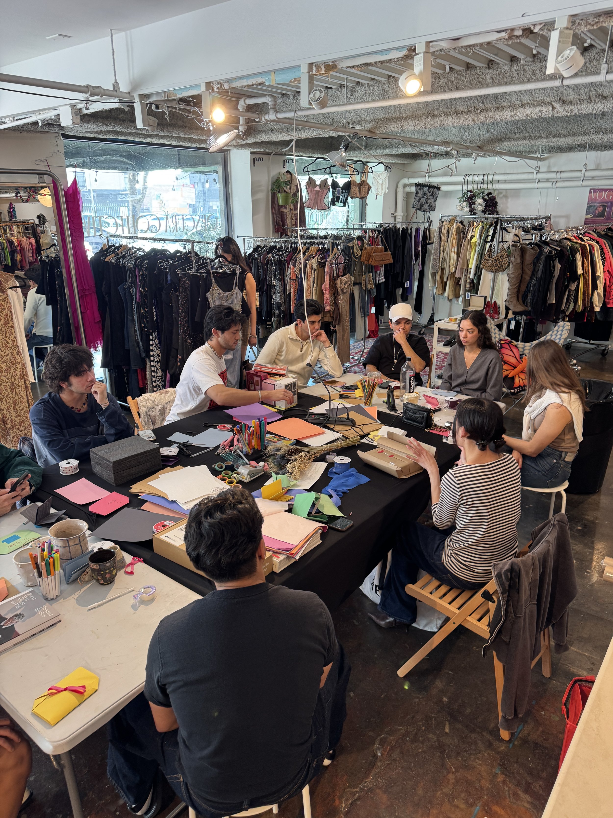 A group of people sitting around a large table in a clothing store, with racks of clothes in the background and crafting supplies on the table.