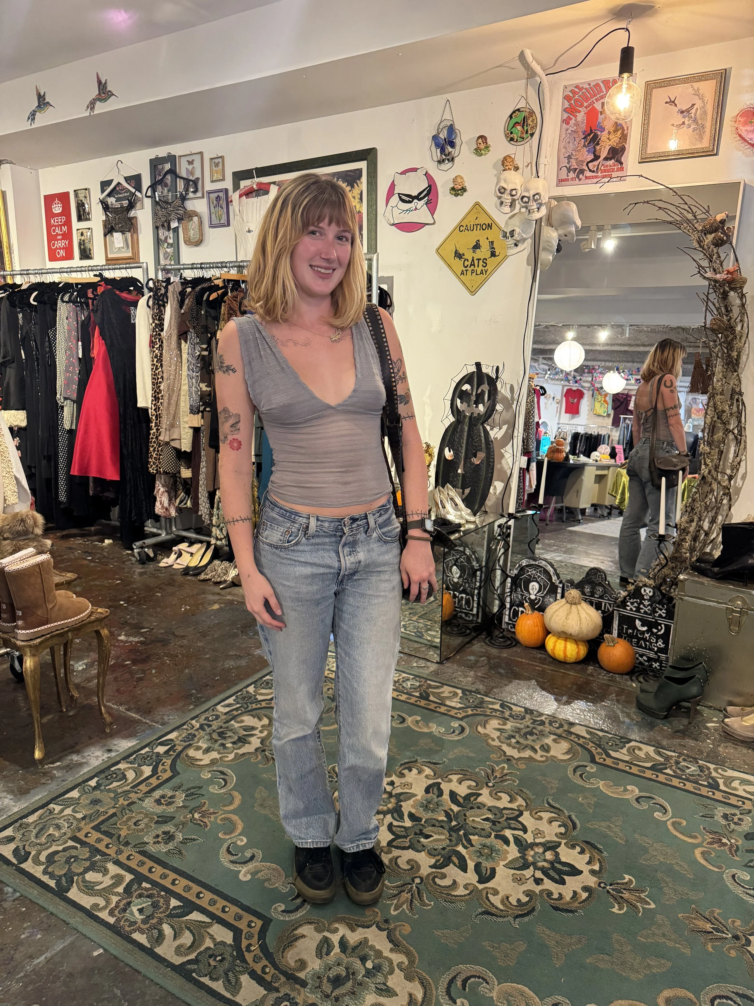 A smiling woman with blonde hair stands in a vintage clothing store, wearing a gray tank top, light blue jeans, and black shoes. The store has various decorations on the wall, holiday pumpkins on the floor, and racks of clothing in the background.