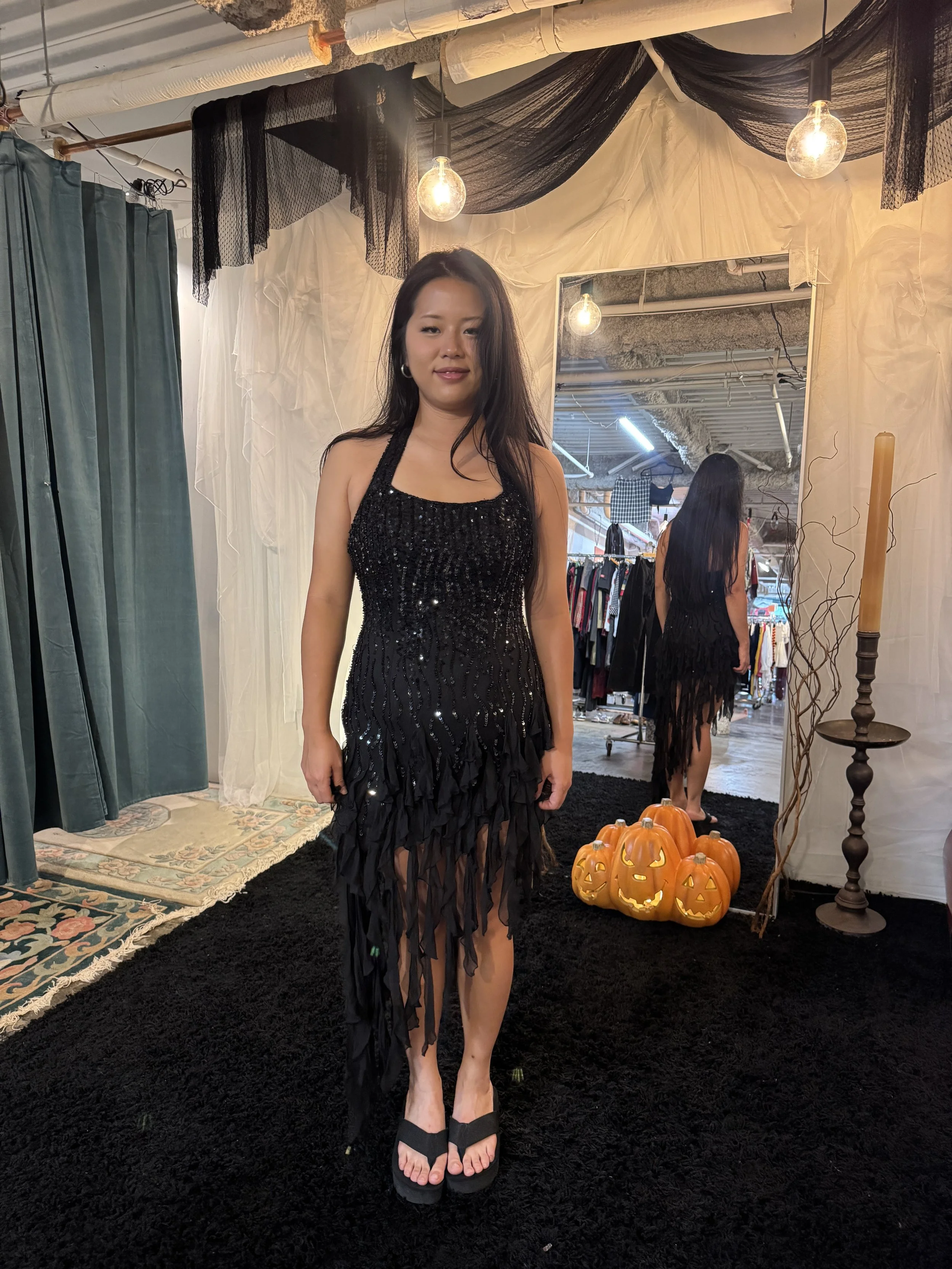A woman in a black, sequin and feather dress standing in front of a mirror in a boutique fitting room, with Halloween pumpkin decorations nearby.