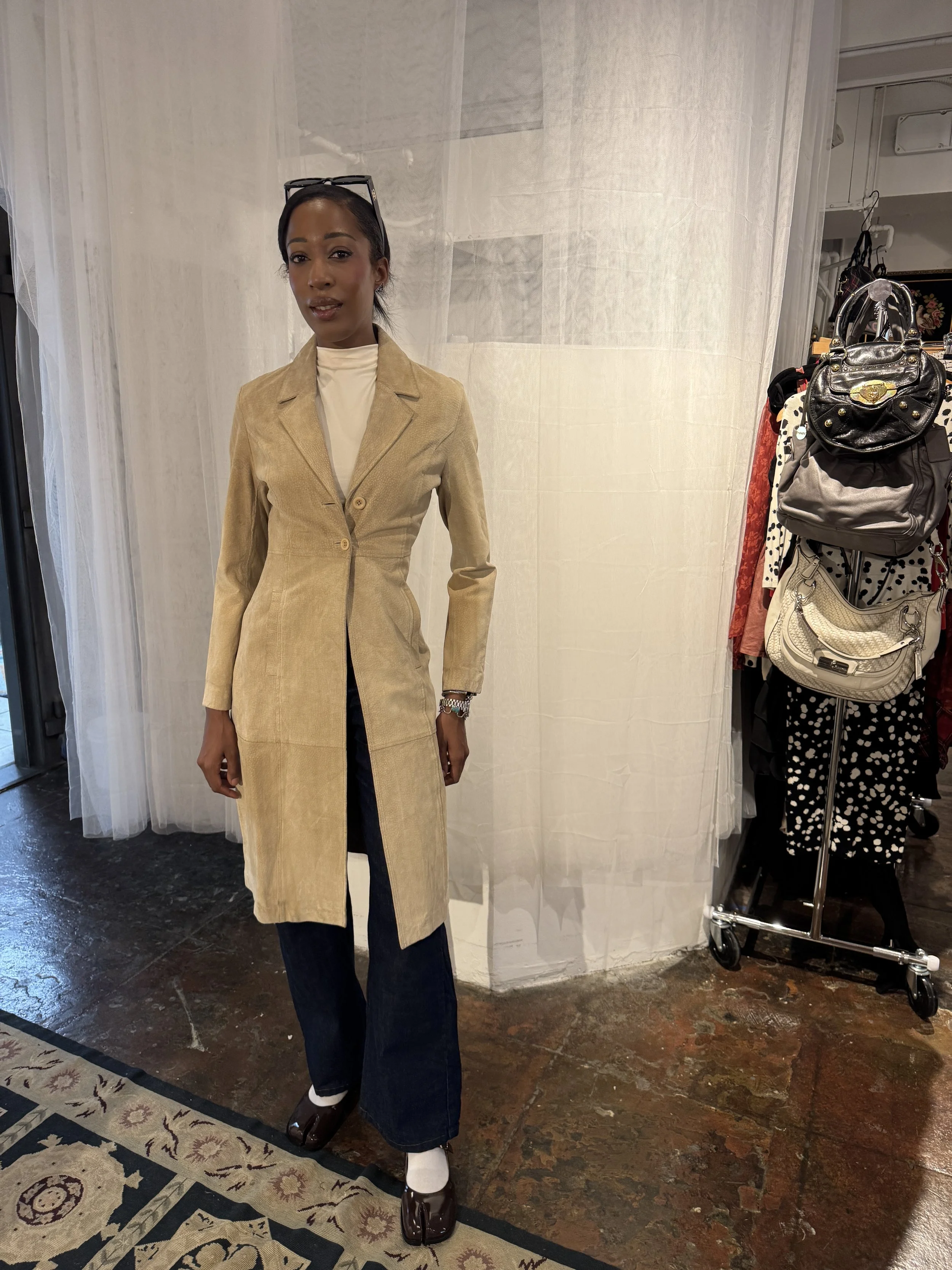 A woman standing indoors wearing a beige long coat, dark flared jeans, and sunglasses on her head, with handbags hanging on a rack nearby.