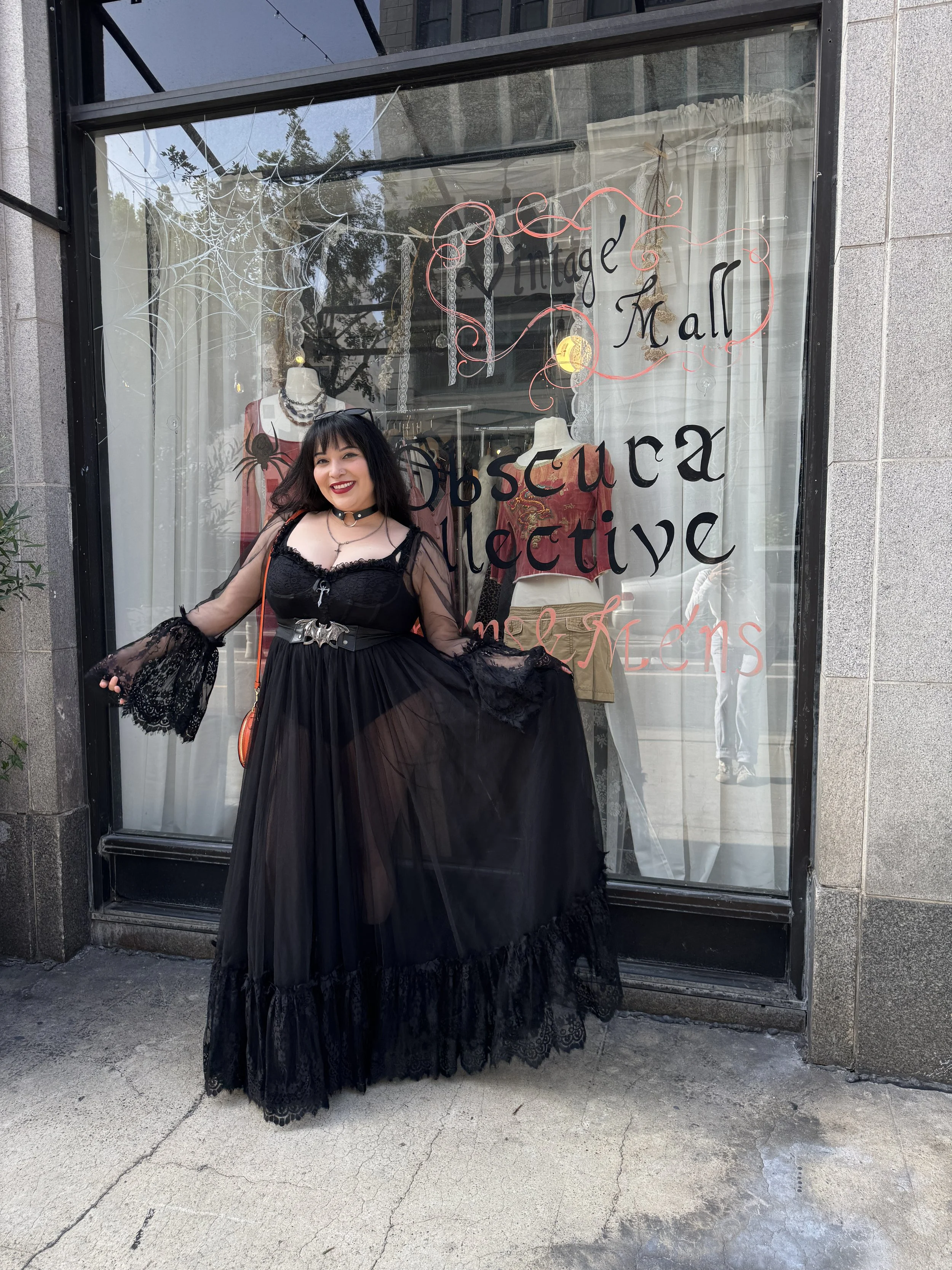 A woman dressed in a black gothic dress with lace sleeves, standing outside an antique and vintage store called 'Obscura Collective.' She is smiling, holding her dress, and wearing makeup with dark lipstick. The store window behind her displays manne