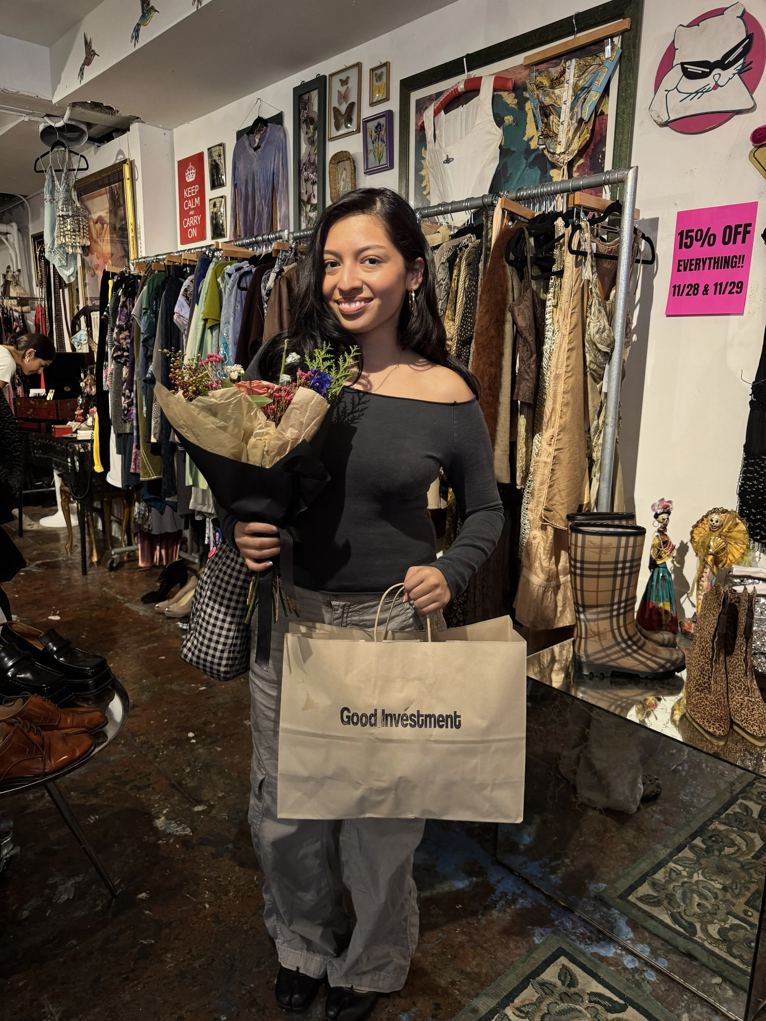 A young woman with dark hair smiling and holding a bouquet of flowers and shopping bag inside a vintage clothing store, surrounded by racks of clothing and accessories.