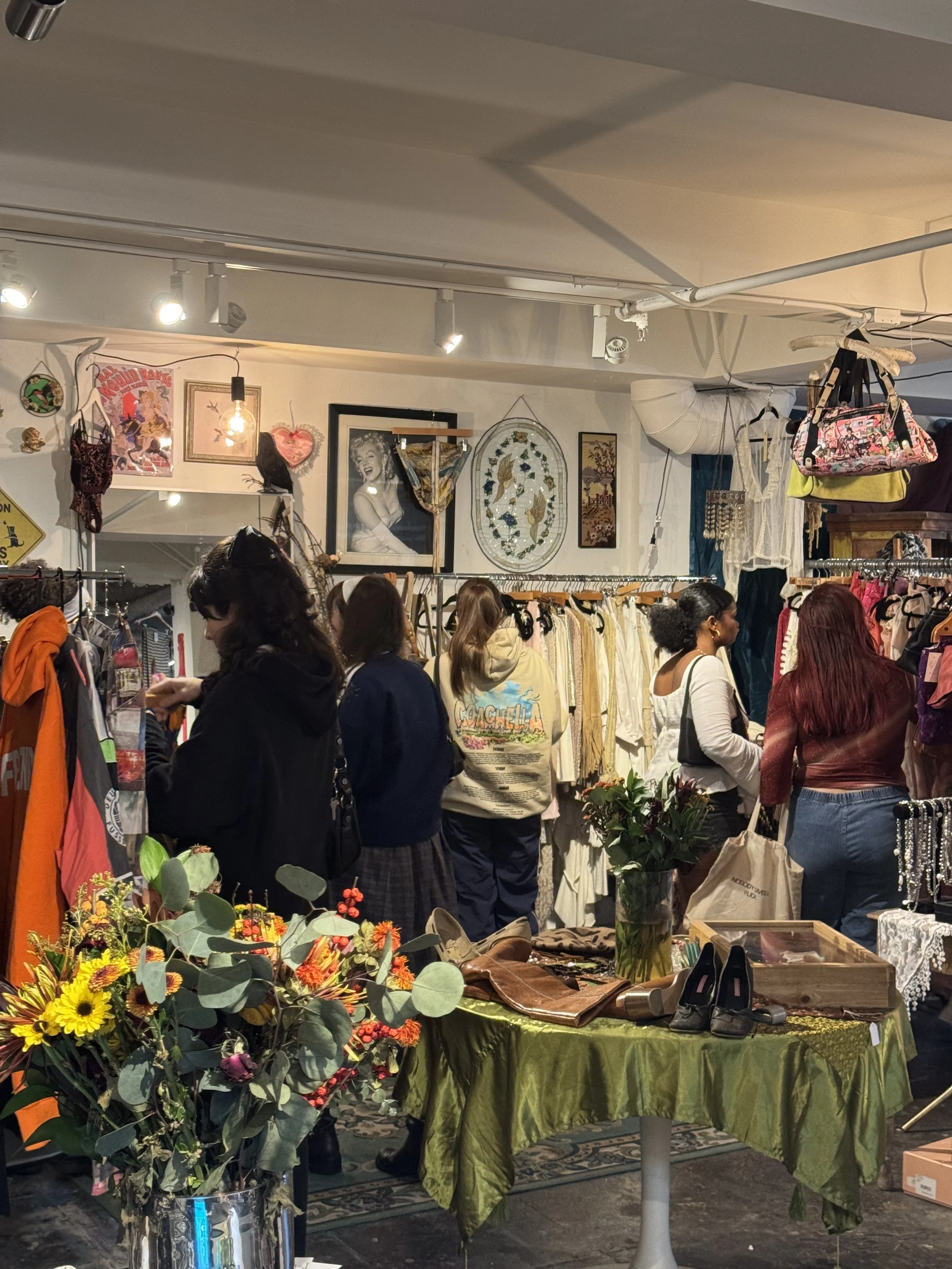 Women shopping at a boutique or thrift store with clothing racks and vintage decor, including framed pictures and decorative wall hangings.