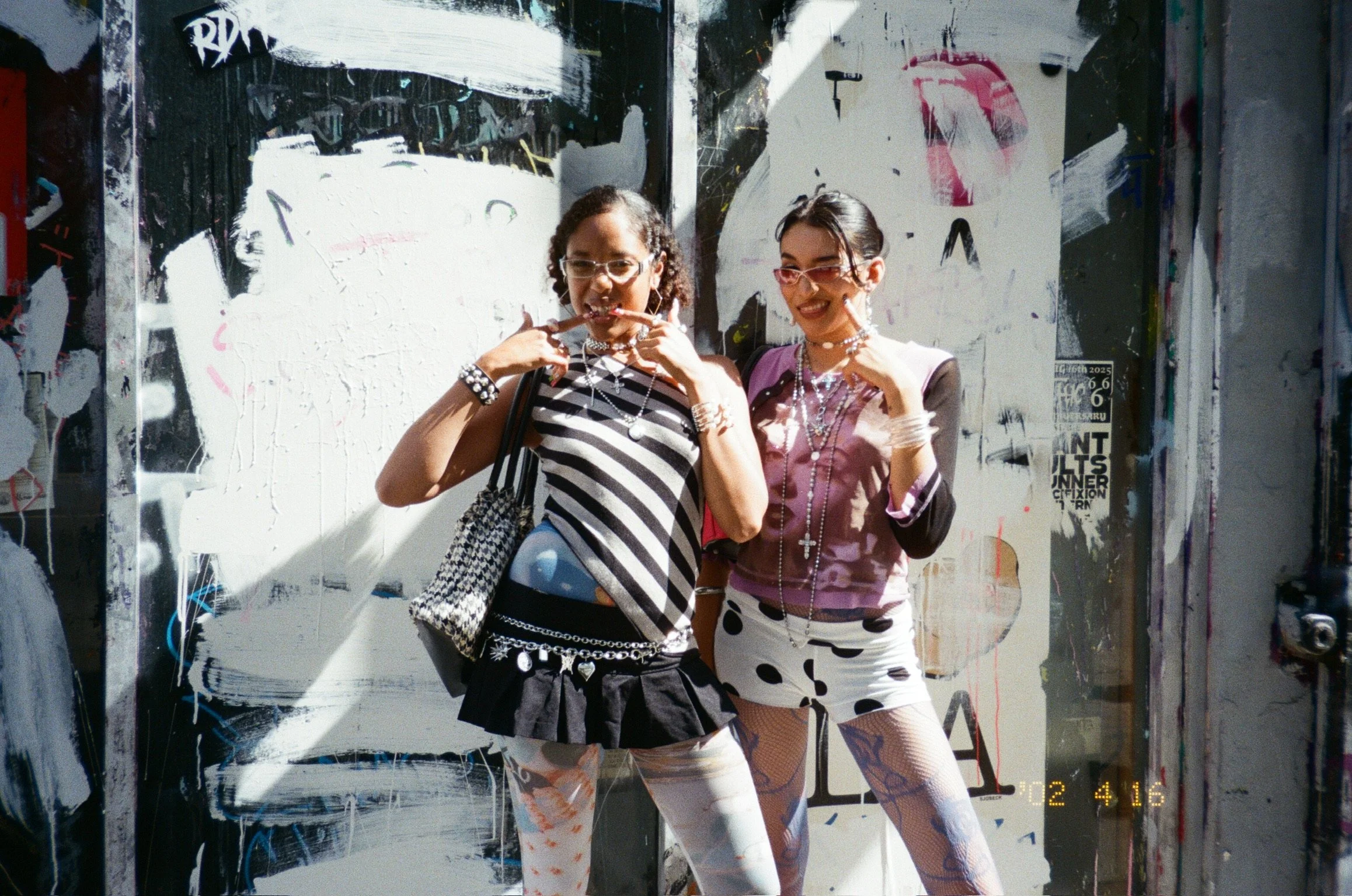 Two women with colorful outfits and accessories standing in front of a graffiti-covered wall, posing playfully.