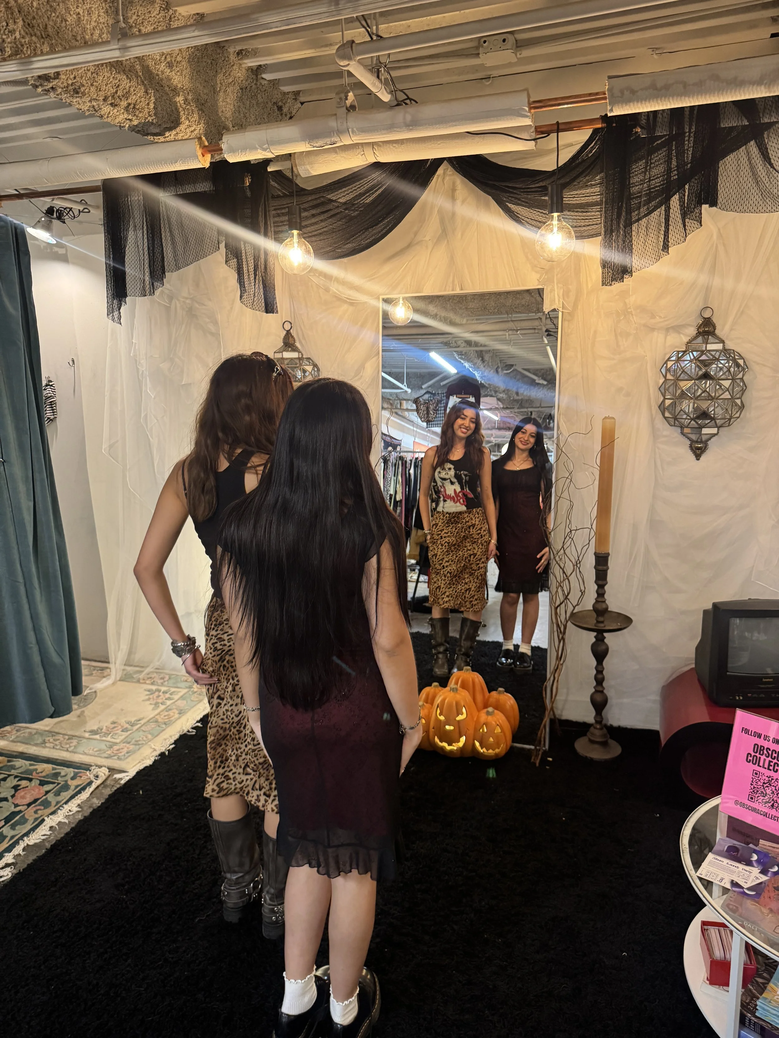 Four women looking at their reflection in a mirror decorated with Halloween-themed pumpkins, lanterns, and drapes in a boutique or dressing room.