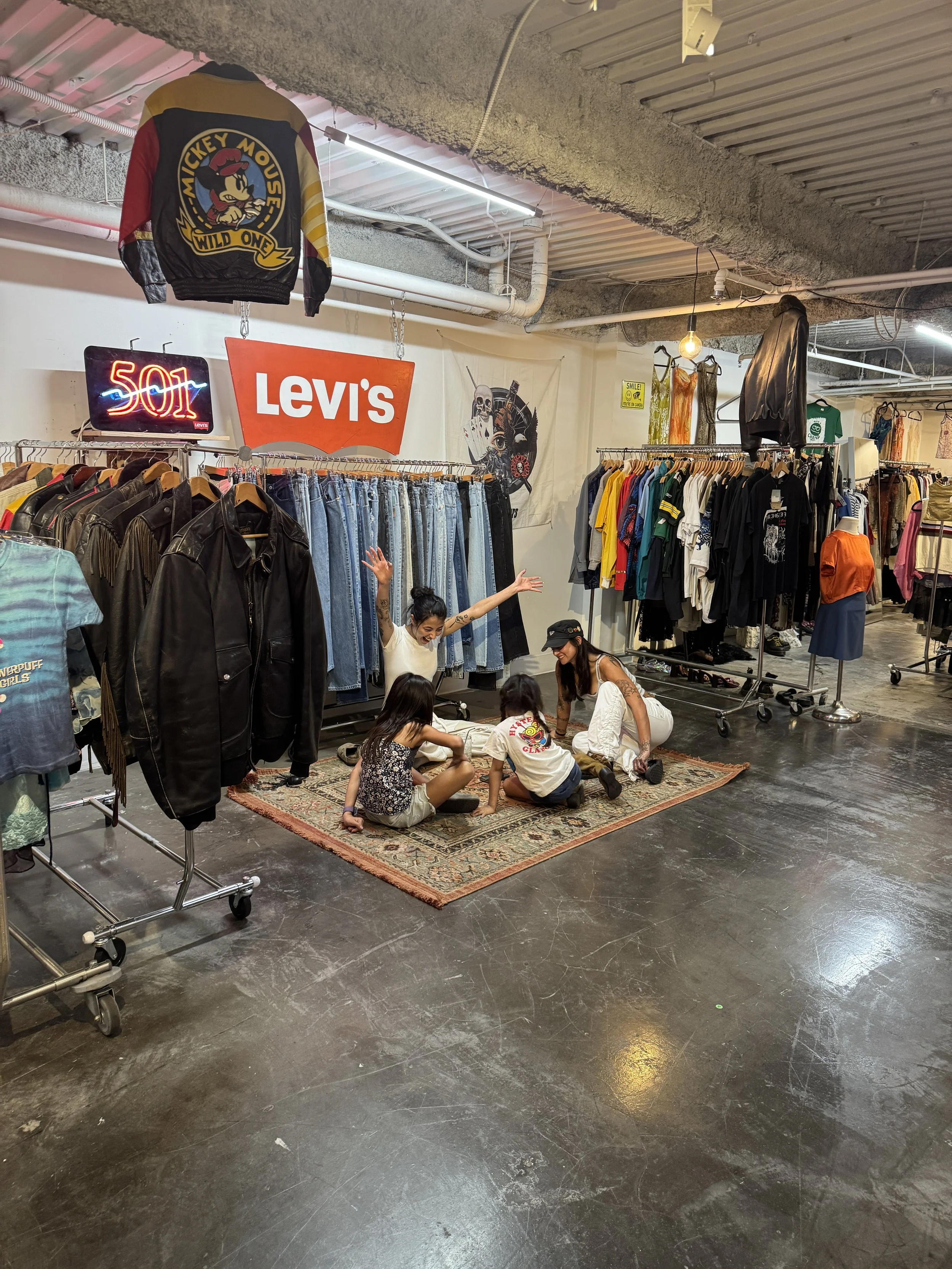 Four children and an adult woman sitting and playing on a small rug inside a vintage clothing store with racks of jackets, jeans, and colorful t-shirts, and vintage signs including a '501' sign and a 'Levi's' sign.