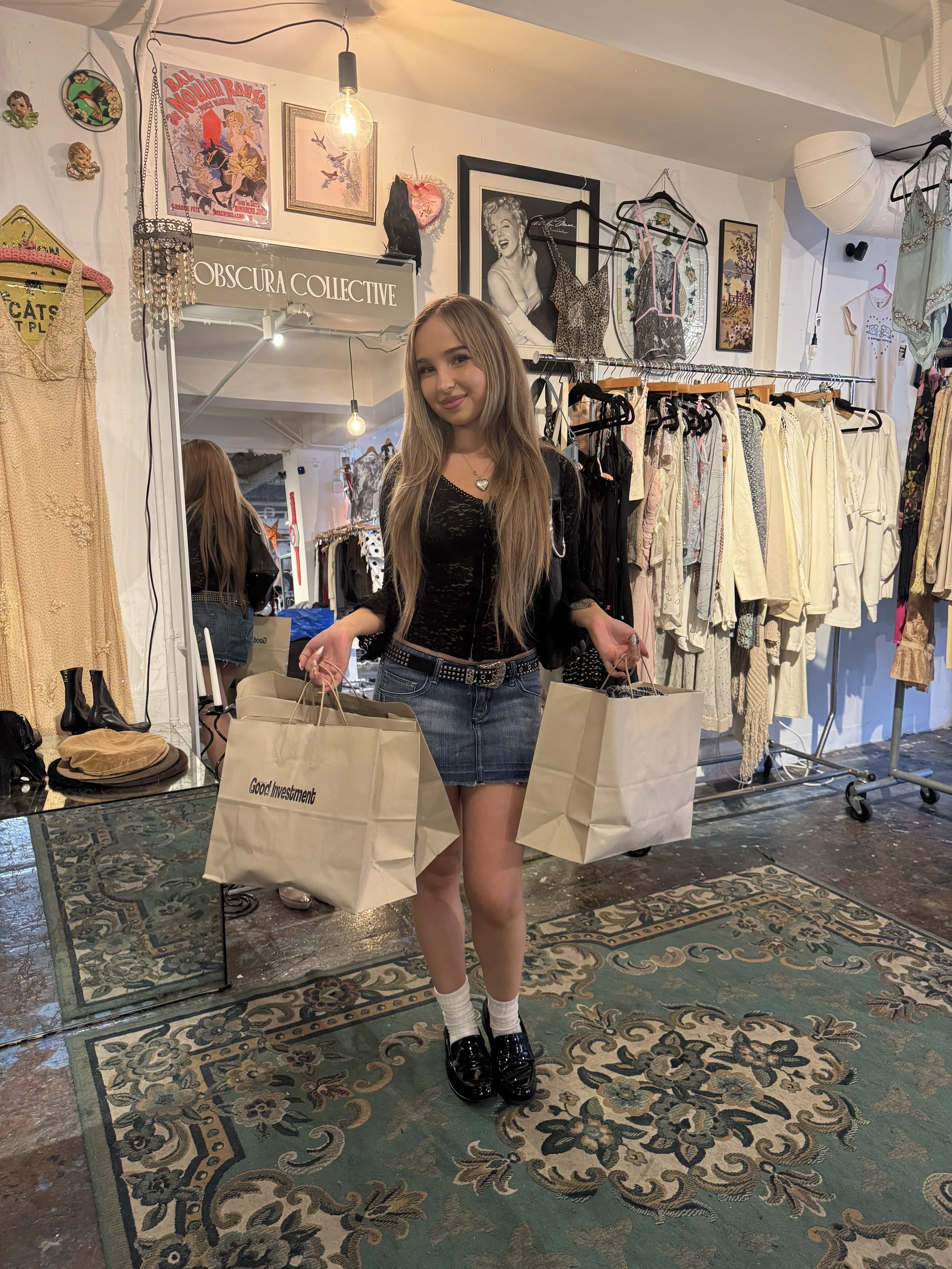 A young woman with long blonde hair holding shopping bags standing in a vintage clothing boutique. The boutique has racks of clothing, framed pictures, and decorative items on the walls.