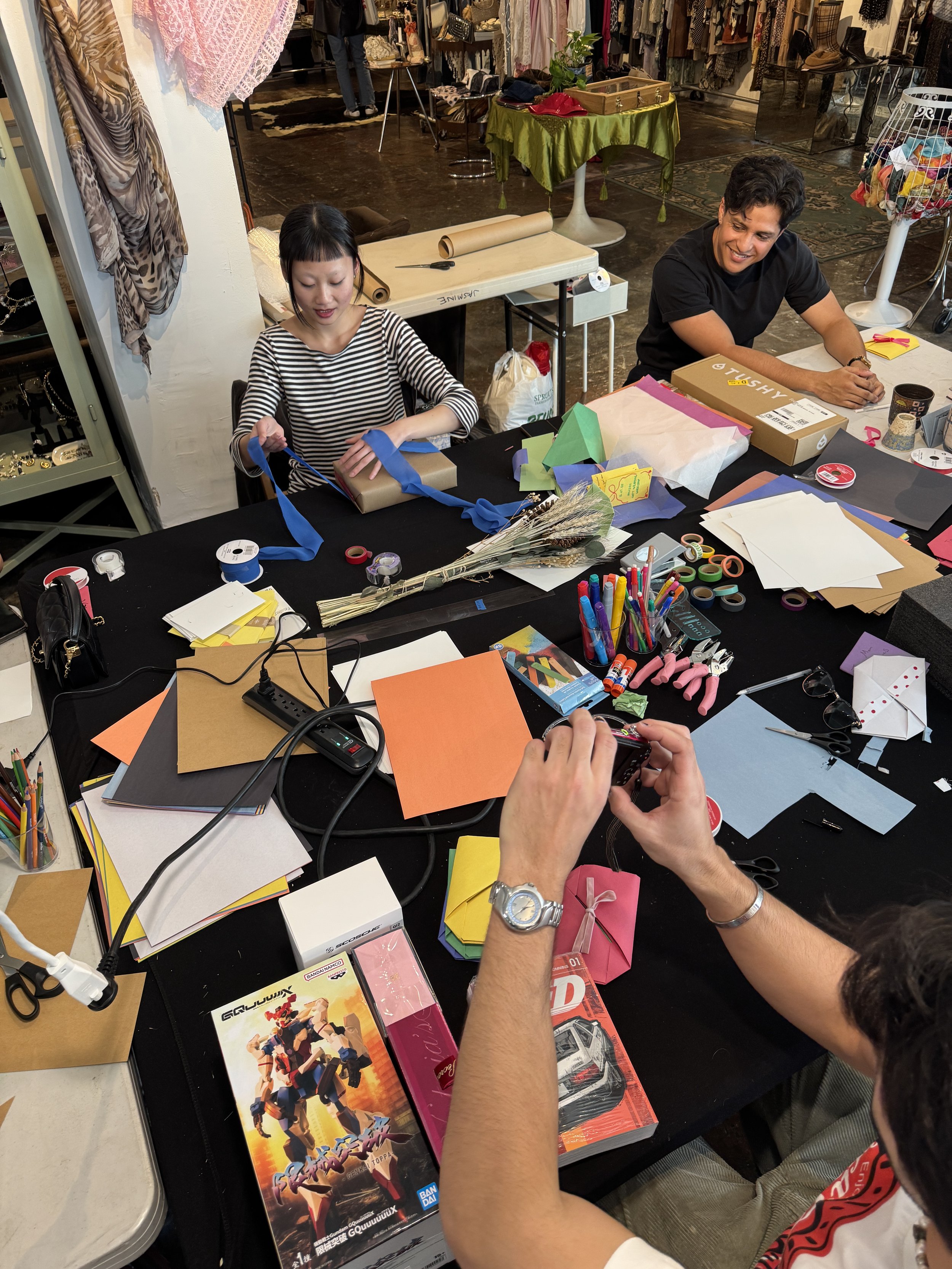 People working on a craft project at a cluttered table with various craft supplies, paper, and scissors in a busy store or studio.