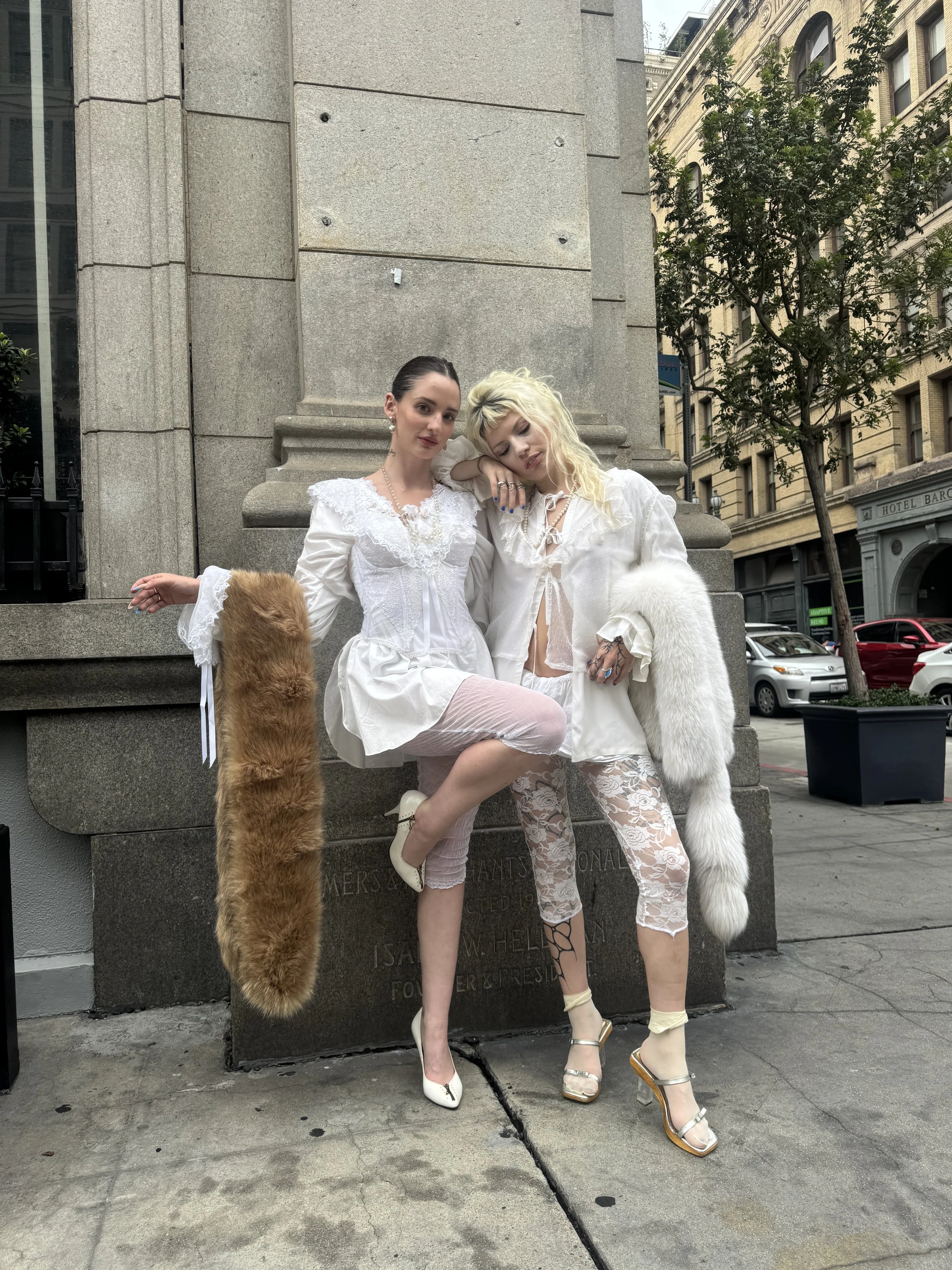 Two women in white outfits posing on a city sidewalk, each wearing fur accessories and high heels.