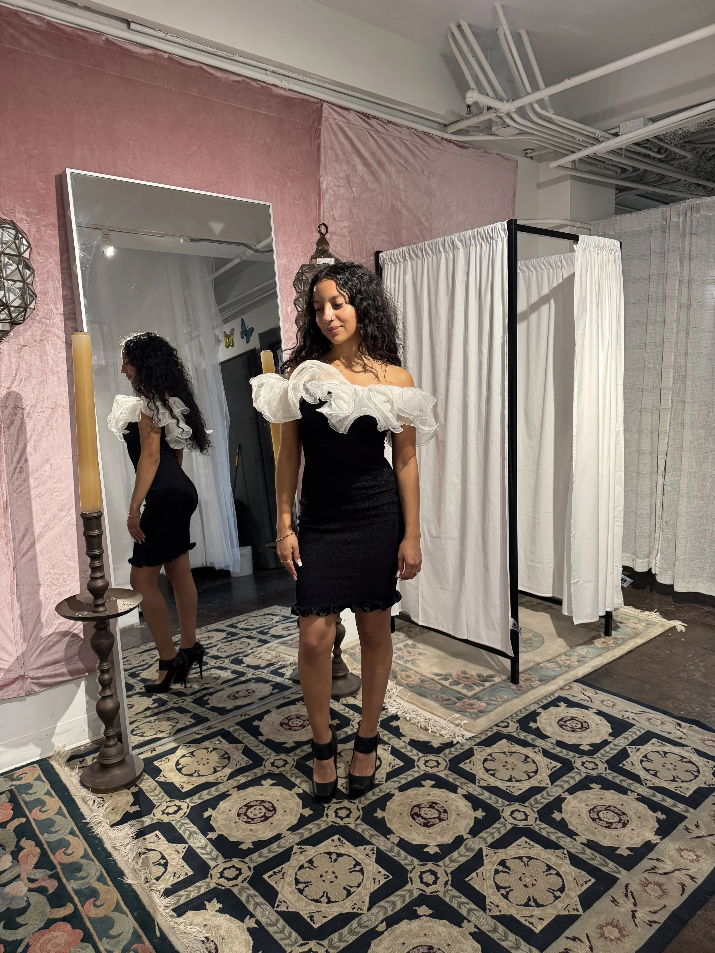 A woman with curly hair wearing a black dress with white ruffled sleeves, black high heels, standing in front of a mirror, with her reflection visible. The setting appears to be a fitting room with a pink wall, a patterned rug, a white curtain, and d
